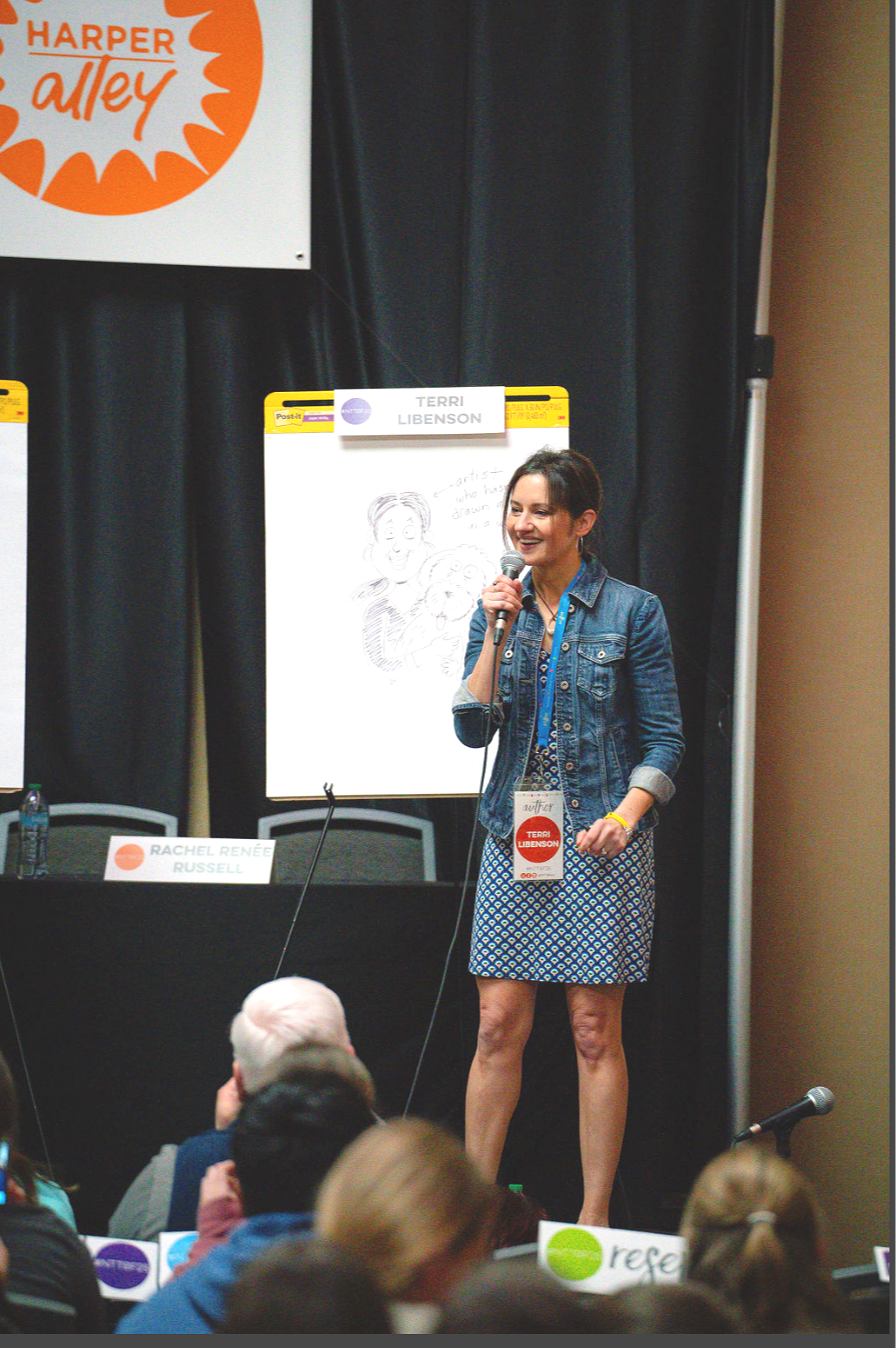 Dallas conference photography of a woman speaking into a microphone on stage at a conference in Dallas, wearing a denim jacket and dress, with a lanyard and badge that says 'Terri Libenson'. Behind her, a large notepad features a cartoon drawing and 