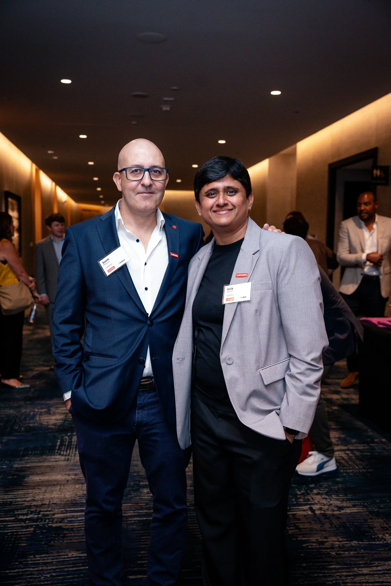 Two men wearing business suits and name tags standing together at an indoor event with other people in the background.