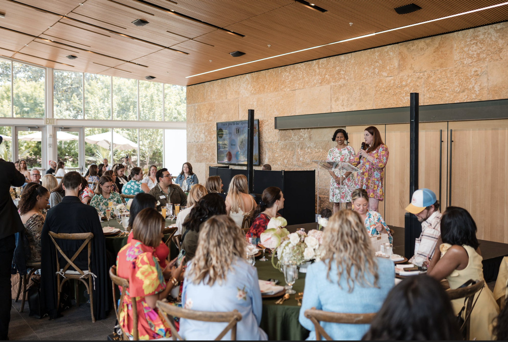 A large indoor gathering with many people seated at round tables listening to two women speaking on a stage. The women are standing at a clear podium, one holding a microphone. The room has large windows with trees outside, and a flat-screen TV displays a slide in the background.