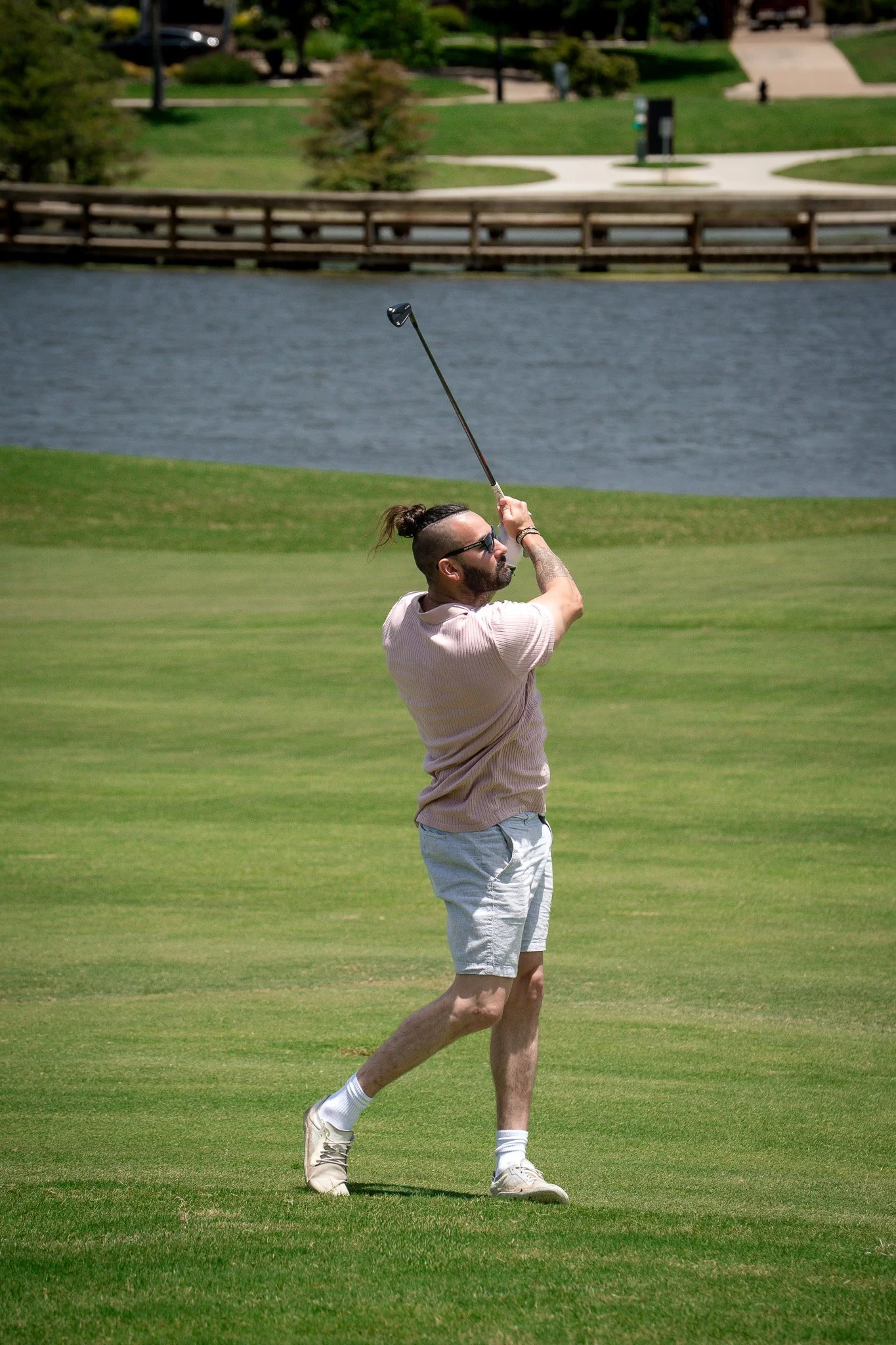 A man playing golf on a green golf course next to a body of water, wearing sunglasses, a light pink shirt, white shorts, and white golf shoes, lifting a golf club after taking a shot.