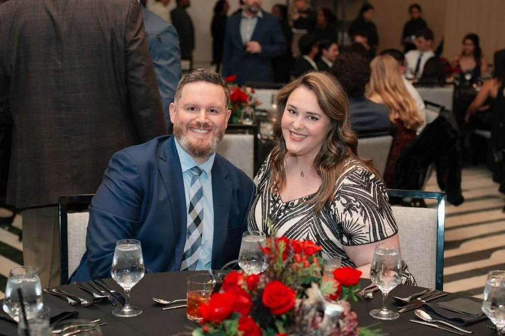 A man and woman smiling at a table during a formal event. The table has glasses and a floral centerpiece with red flowers.
