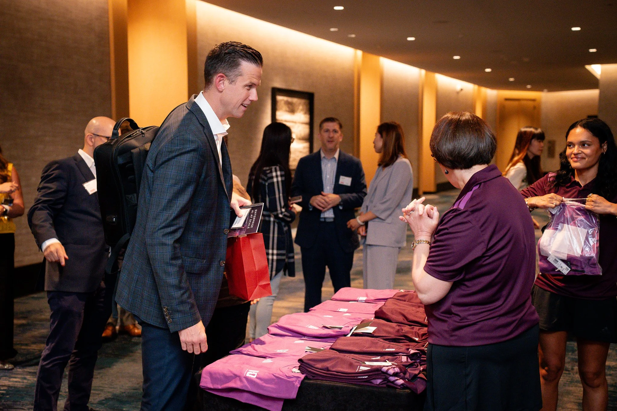 A business conference or event with people interacting at a registration or merchandise table, with a man in a checkered suit talking to a woman at the table.