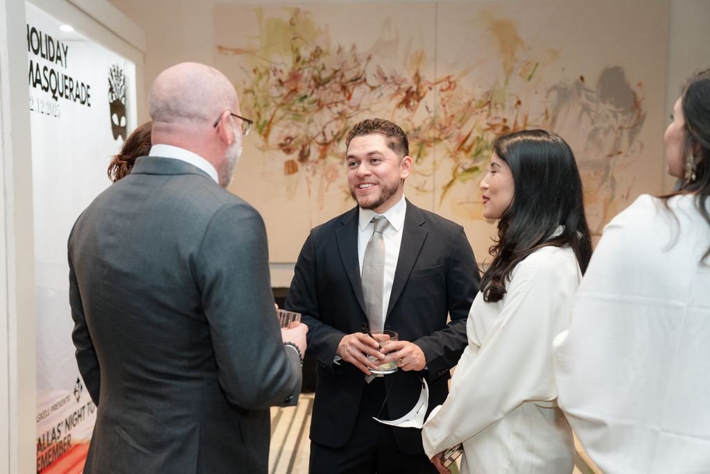 Group of four people in formal attire conversing at an indoor event, smiling and holding drinks.
