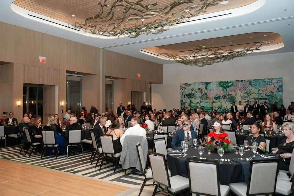 Guests seated at banquet tables in a large decorated event hall, with a modern ceiling design and artwork on the back wall.