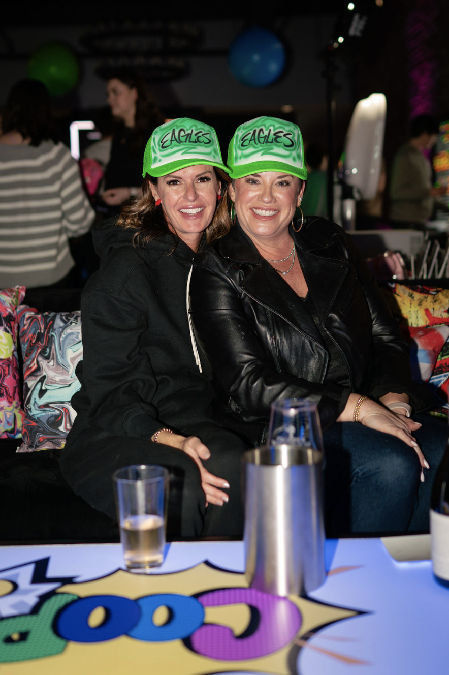Two women sitting together at a party, both wearing bright green hats with 'EAGLES' written on them, smiling at the camera. In the background, other party guests and colorful balloons can be seen.