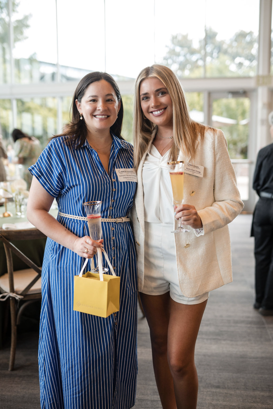 Two women standing side by side at an indoor event, smiling and holding champagne glasses, with name tags on their outfits.