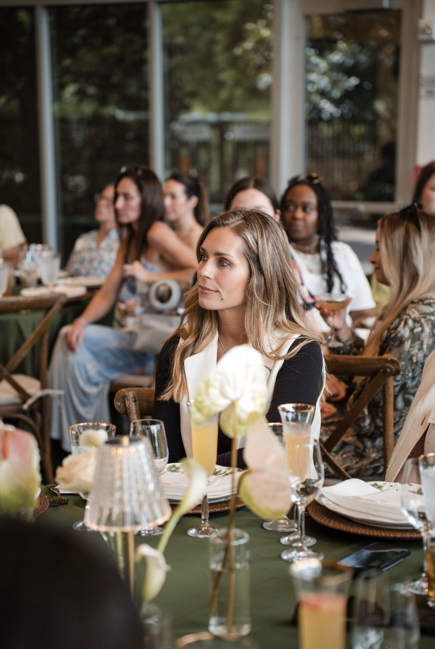 A woman with long blonde hair wearing a black and white outfit sitting at a decorated table during a gathering or event, with other women in the background.