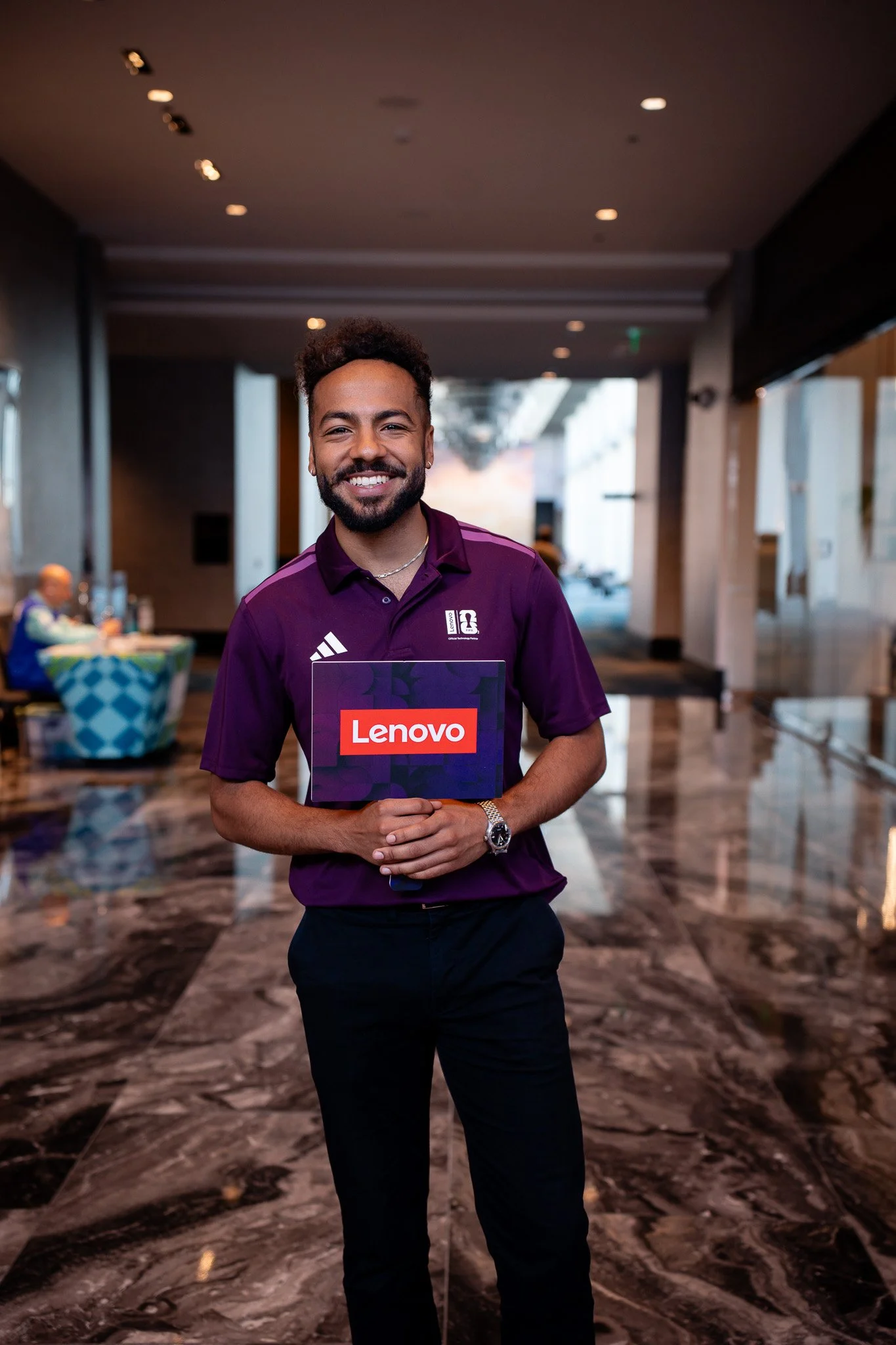 Smiling man in purple shirt holding a Lenovo sign, standing in a hotel lobby with marble floors and blurred background of people and decor.