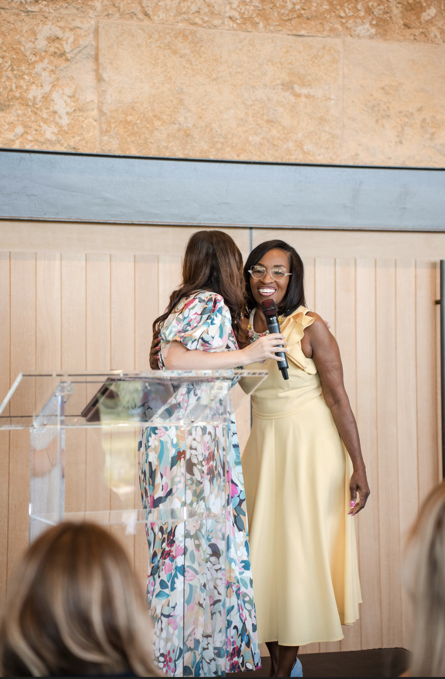 Two women sharing a microphone and smiling on stage, one in a floral dress and the other in a yellow dress, during an indoor event.