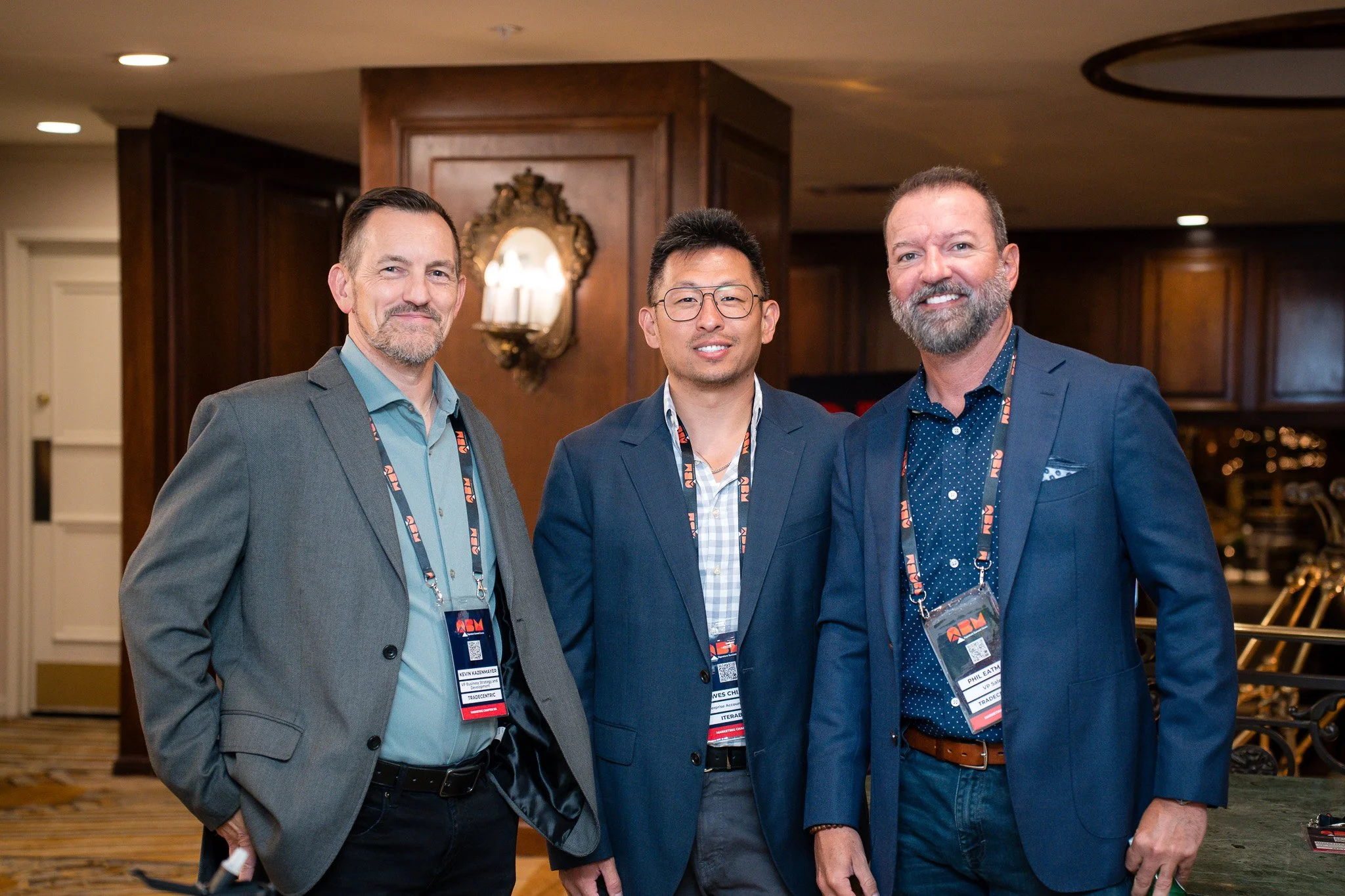 Three men in suits standing together at a conference, smiling for the camera, indoors with warm lighting and wooden decor in the background.
