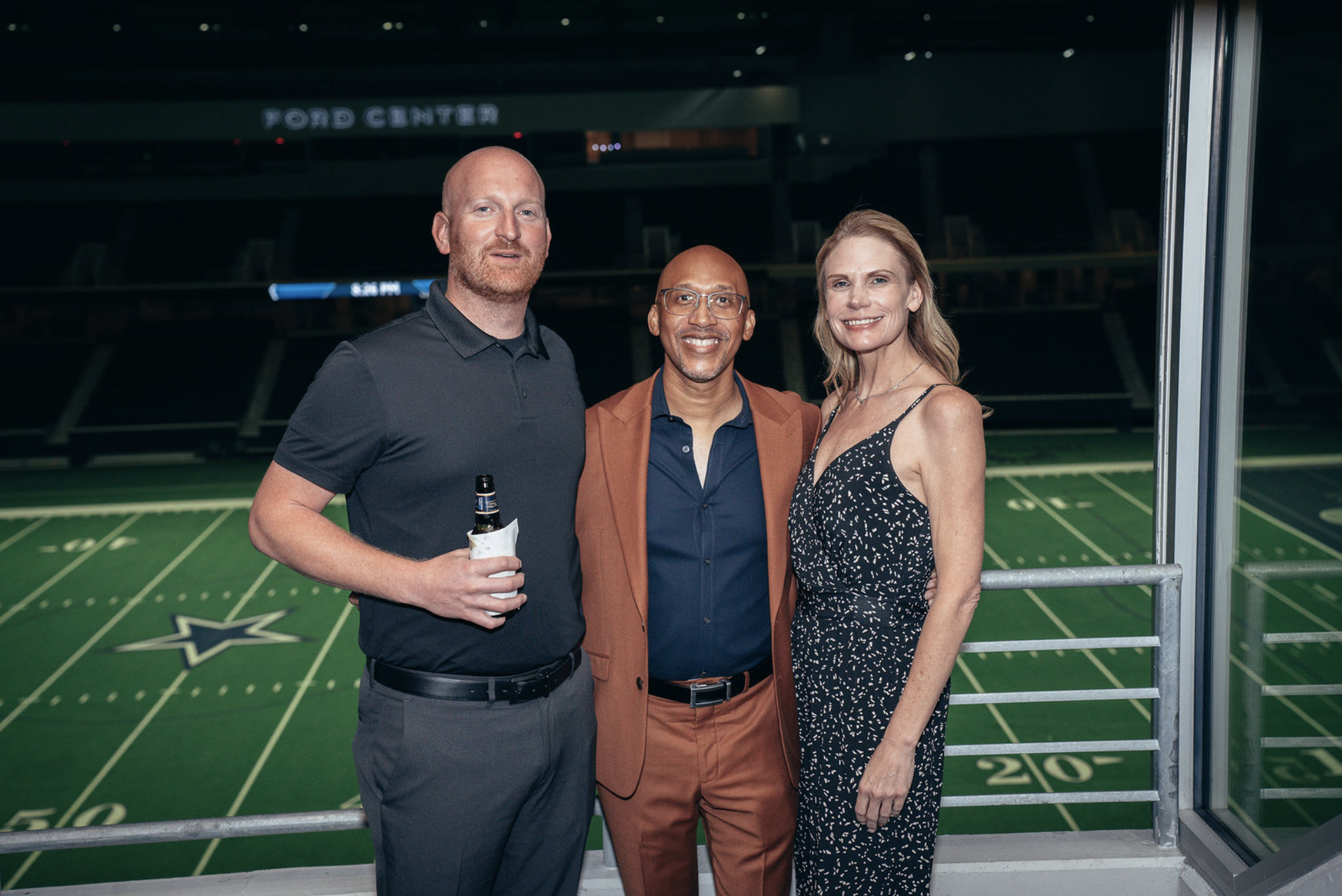 Three people standing on a balcony at AT&T Stadium, home of the Dallas Cowboys, with a football field in the background. One man is holding a beer, and all three are smiling.