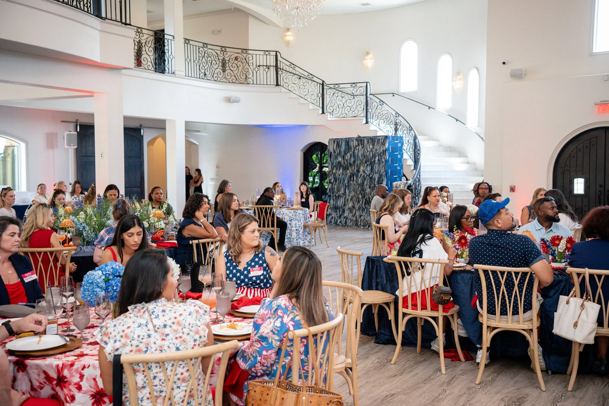 A large indoor event space filled with diverse people sitting at tables, attending a gathering or celebration with floral decorations and table settings, high ceilings, ornate staircases, chandeliers, and large windows.