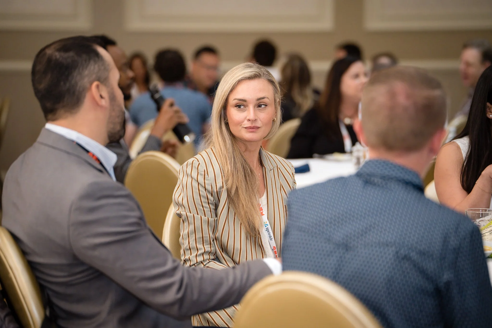 A woman with blonde hair and a striped blazer sits at a conference table, looking to her left during a professional event in a conference room filled with attendees.