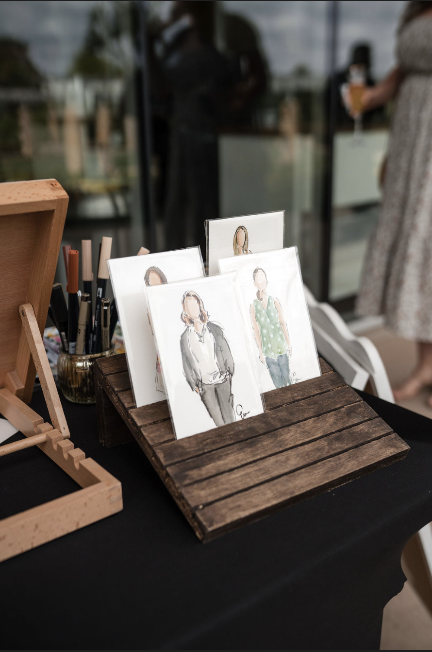 Watercolor portraits of women displayed on a wooden stand at an art fair, with markers in a holder nearby.