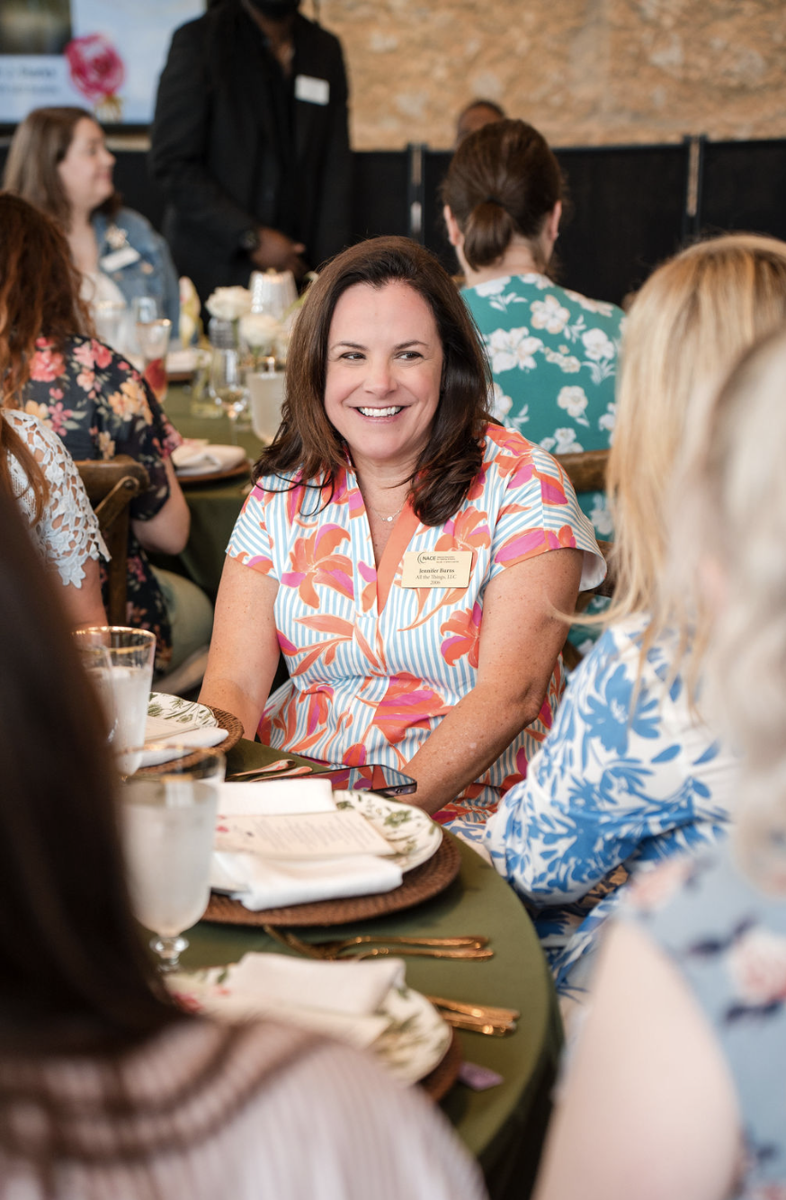 A woman at a formal event or conference, wearing a colorful, floral-patterned dress, smiling and engaging in conversation with others at a round table set with dinnerware and glasses.