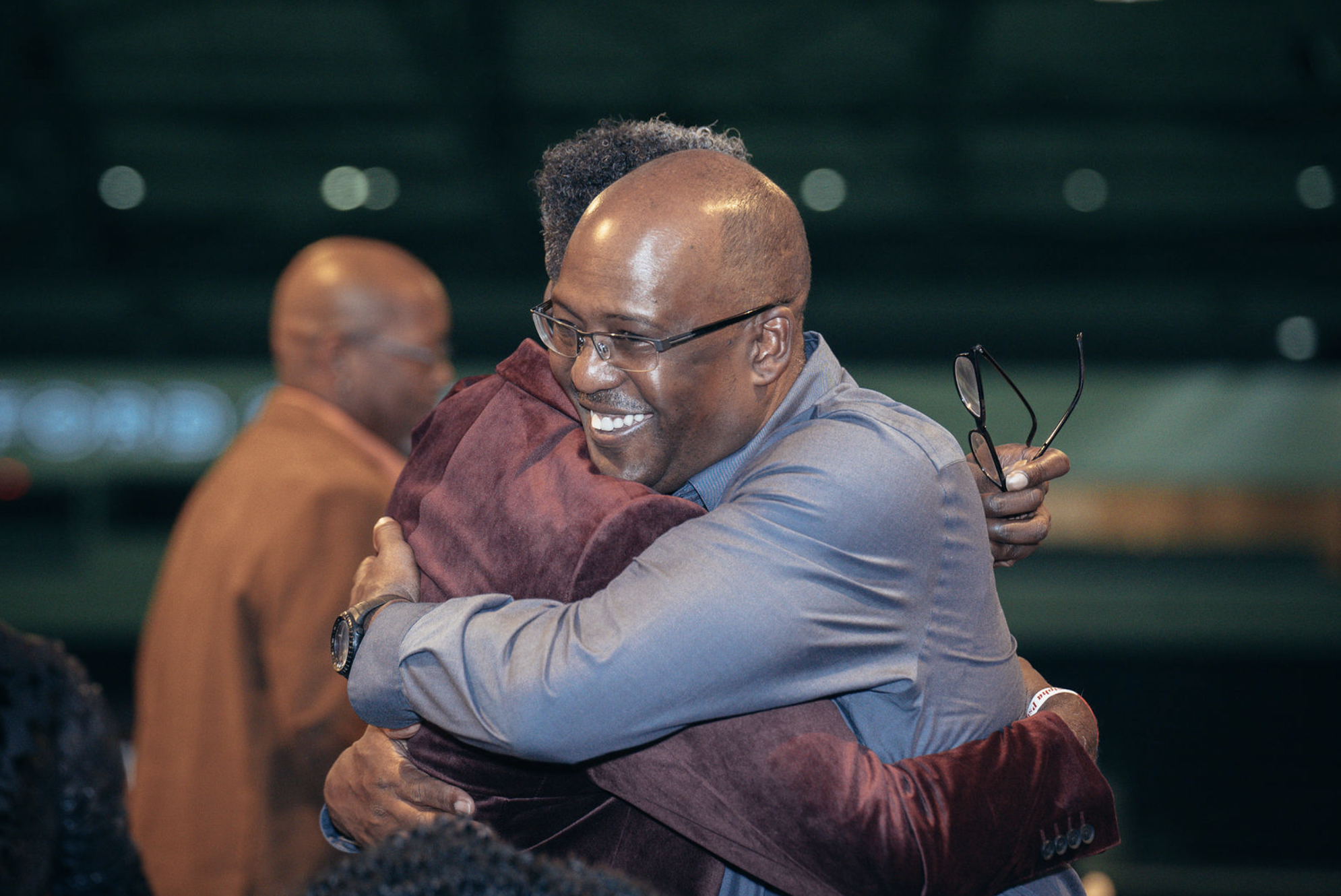 Two men hugging, one smiling and holding glasses, in an indoor setting with other blurred people in the background.