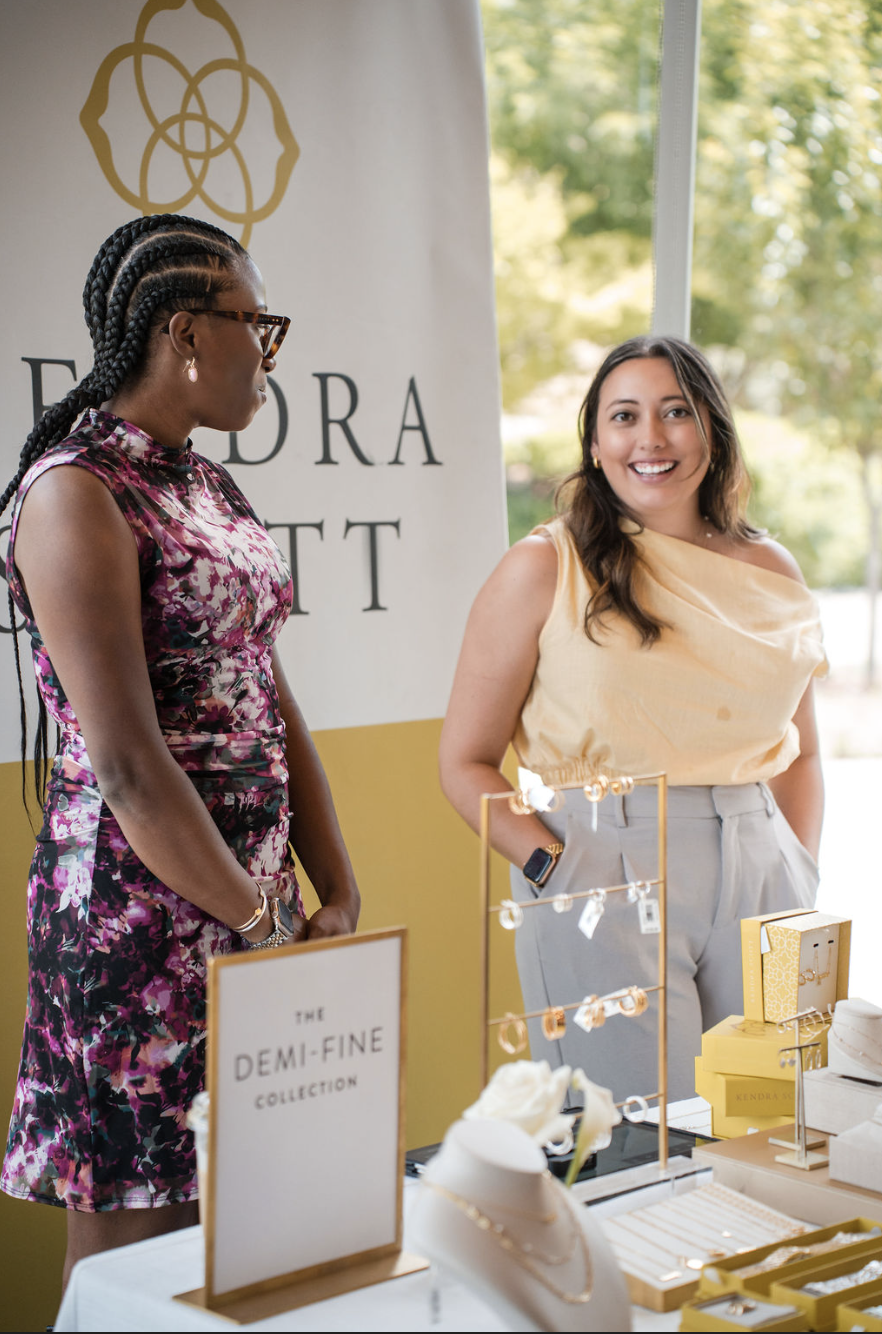 Two women talking at a jewelry booth with a sign that reads 'The Demi-Fine Collection'; one woman is wearing a floral dress and glasses, the other is in a yellow top and gray pants, smiling. The booth displays jewelry and accessories.