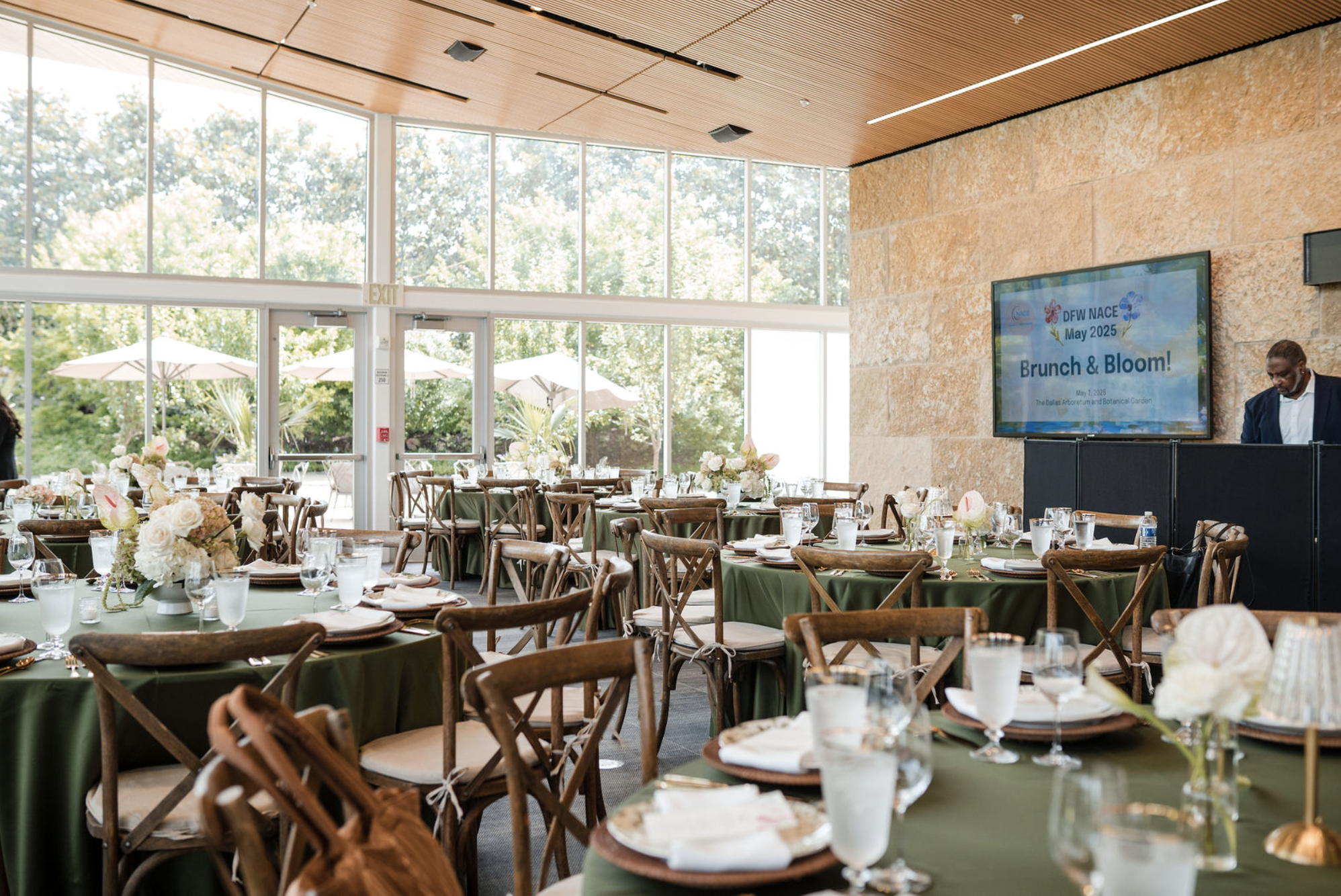 Event setup in a bright, modern indoor space with round tables covered in green tablecloths, decorated with floral centerpieces, set with glassware, plates, and napkins. Outside, patio tables with umbrellas are visible through large glass windows. A large screen displays information about a brunch event titled 'Brunch & Bloom!' scheduled for May 1, 2025.