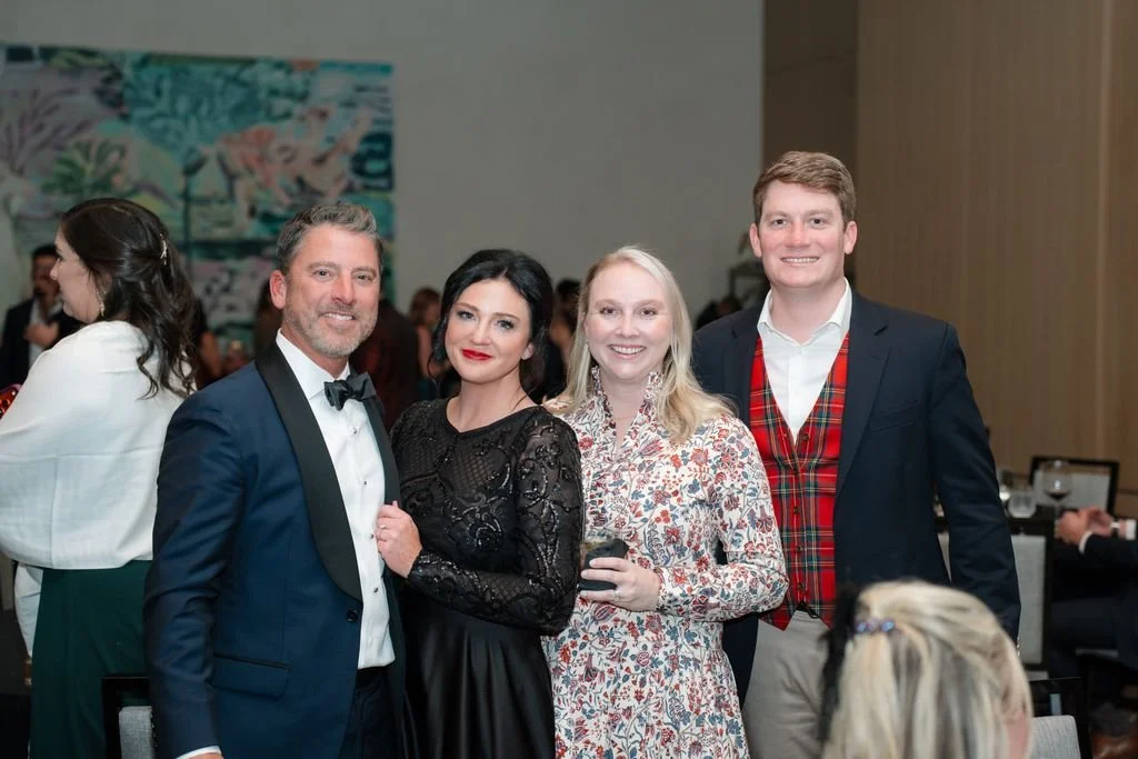 Four people at a formal event, dressed in evening attire, posing for a group photo, with a blurred background of other guests and artwork on the wall.