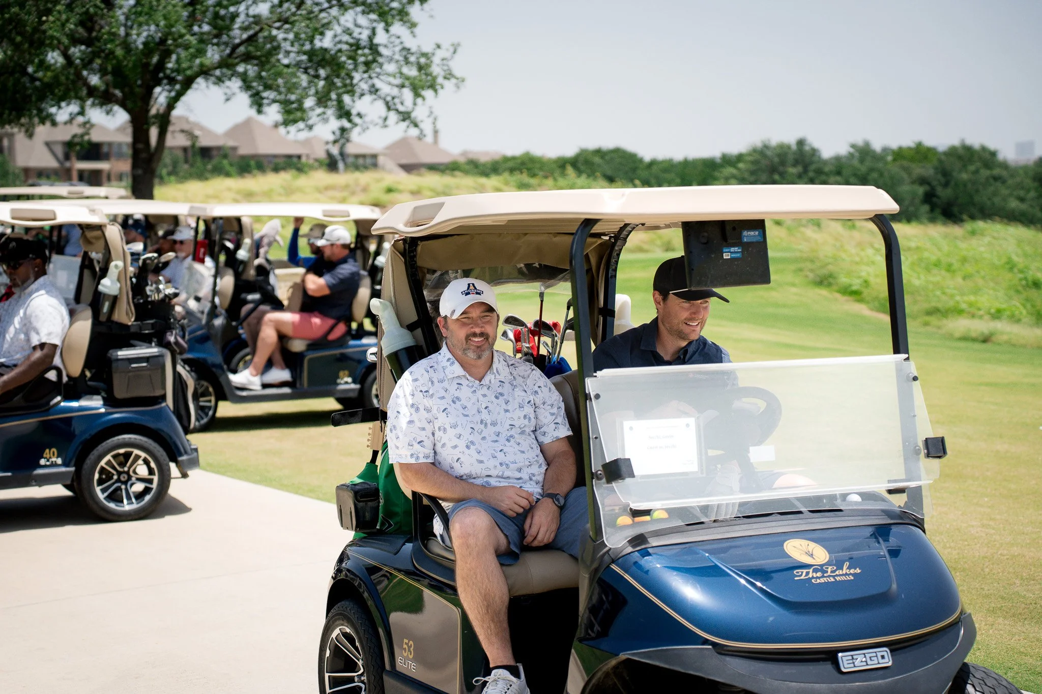 A group of people sitting in golf carts on a golf course, with two men in the foreground smiling, one wearing a hat with the 'Andrews' logo. The background shows more golf carts and trees under a partly cloudy sky.