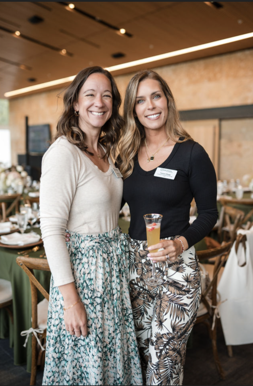 Two women at a social event standing close together and smiling at the camera, one holding a drink, in a decorated event space with tables and chairs.