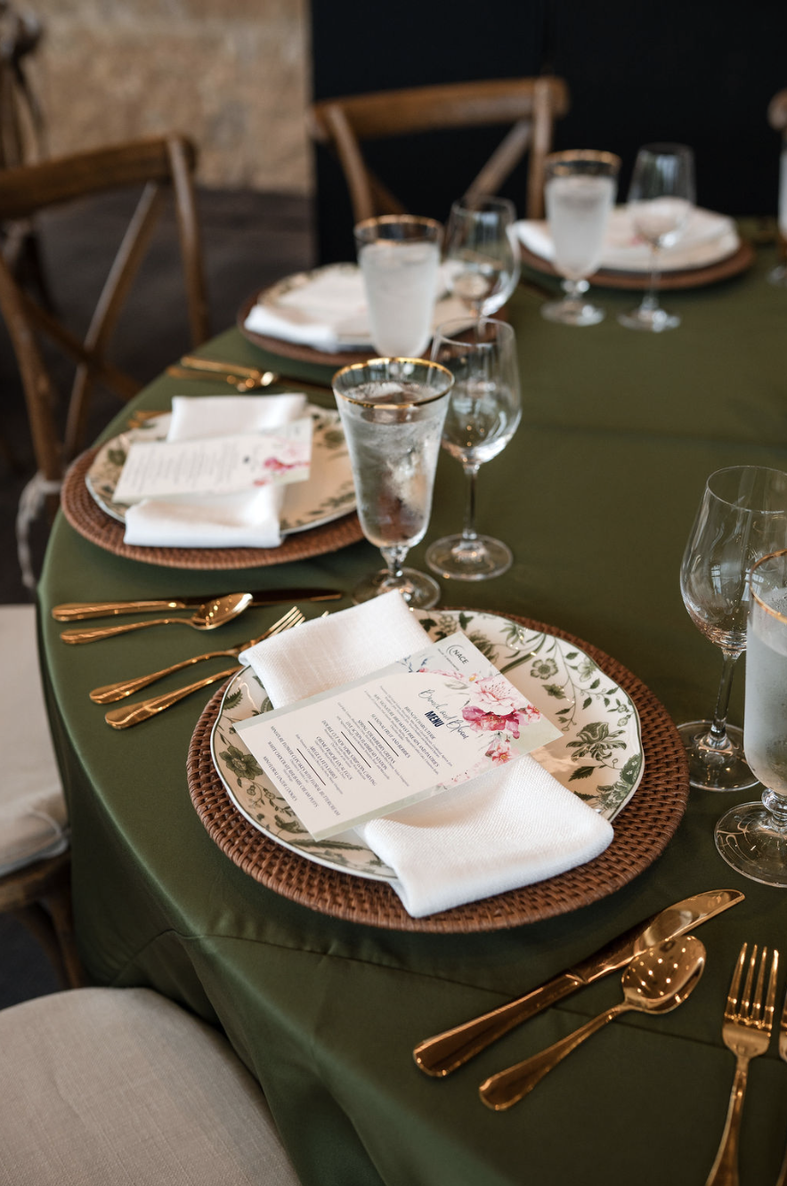 A formal dining table set with floral-patterned plates, white napkins, gold utensils, and glasses of water, with a green tablecloth and wooden chairs surrounding it.