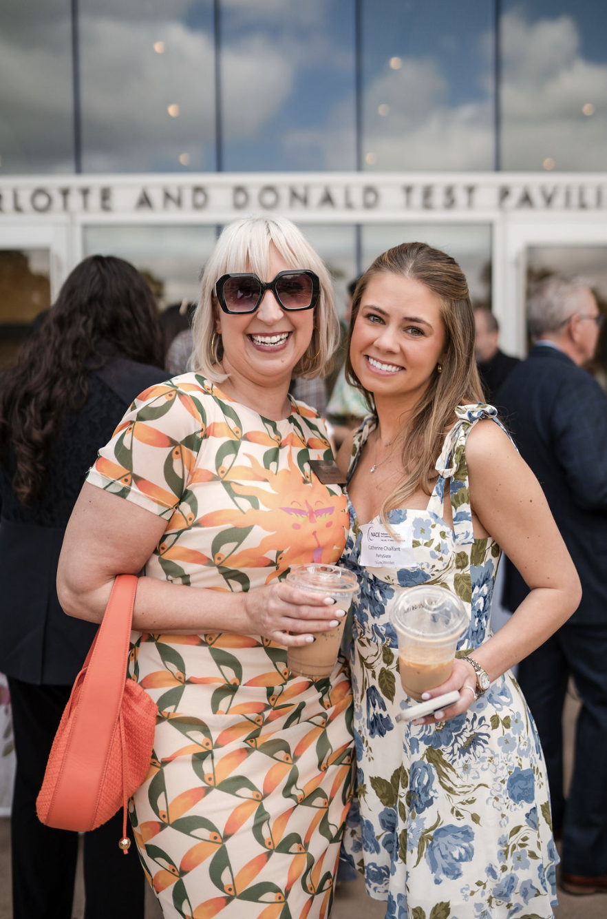 Two women at an outdoor event smiling and holding drinks, standing in front of a building with glass windows and a sign that reads 'Loyola and Donald Test Pavilion.'