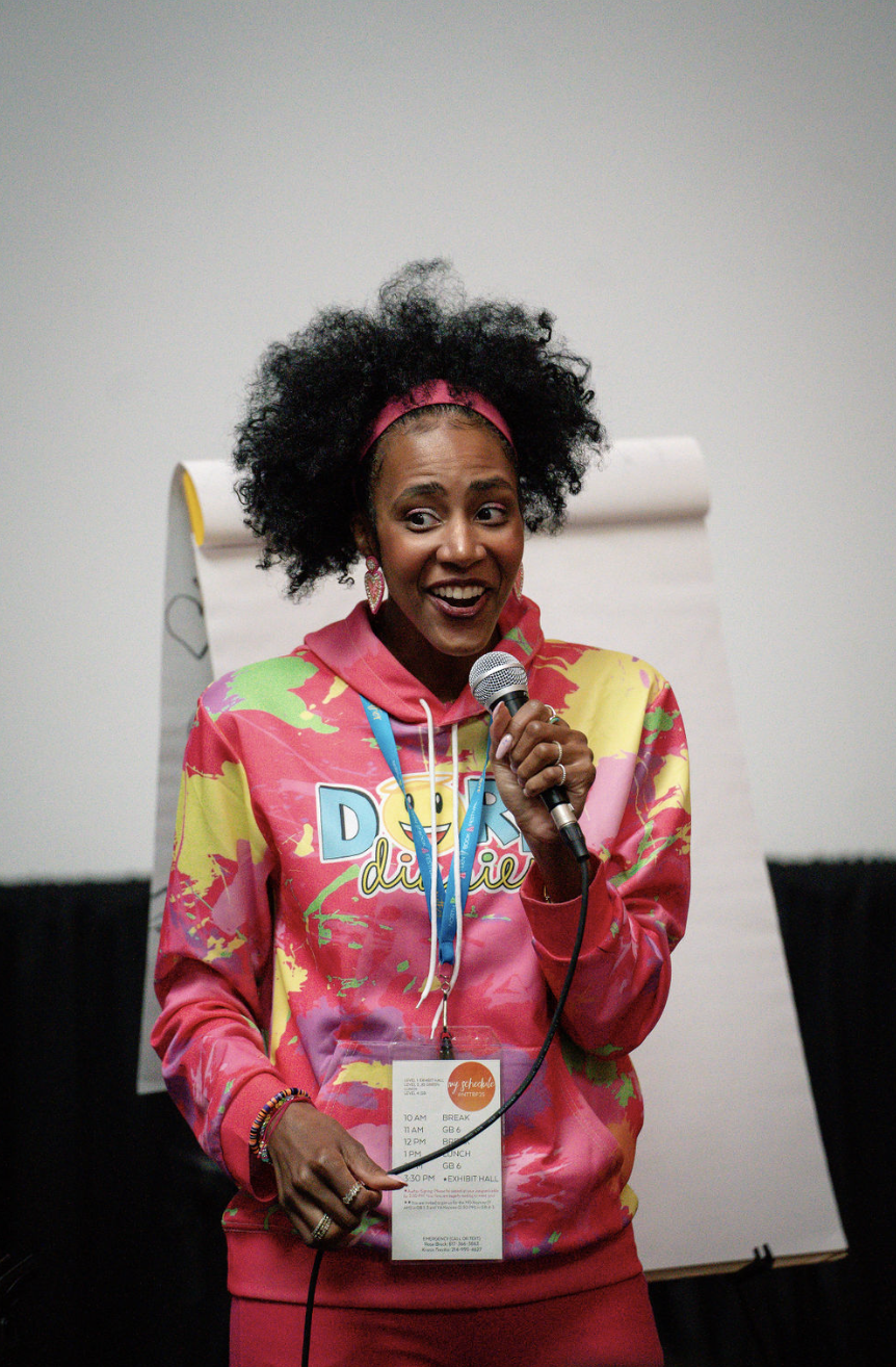 A woman with large curly hair, wearing a pink hoodie with colorful patterns and text, holding a microphone, and smiling during a presentation or speech.