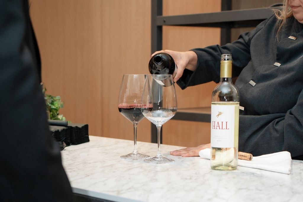 Person pouring white wine into a glass at a marble counter, with another glass of red wine already filled, a bottle of wine labeled 'HALL', and a corkscrew.