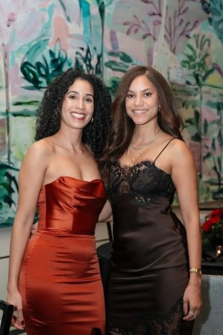 Two women in elegant dresses standing close together indoors. One wears a strapless, shiny red dress, and the other a black dress with lace details. Both are smiling at the camera, with a colorful abstract mural and some red flowers in the background.