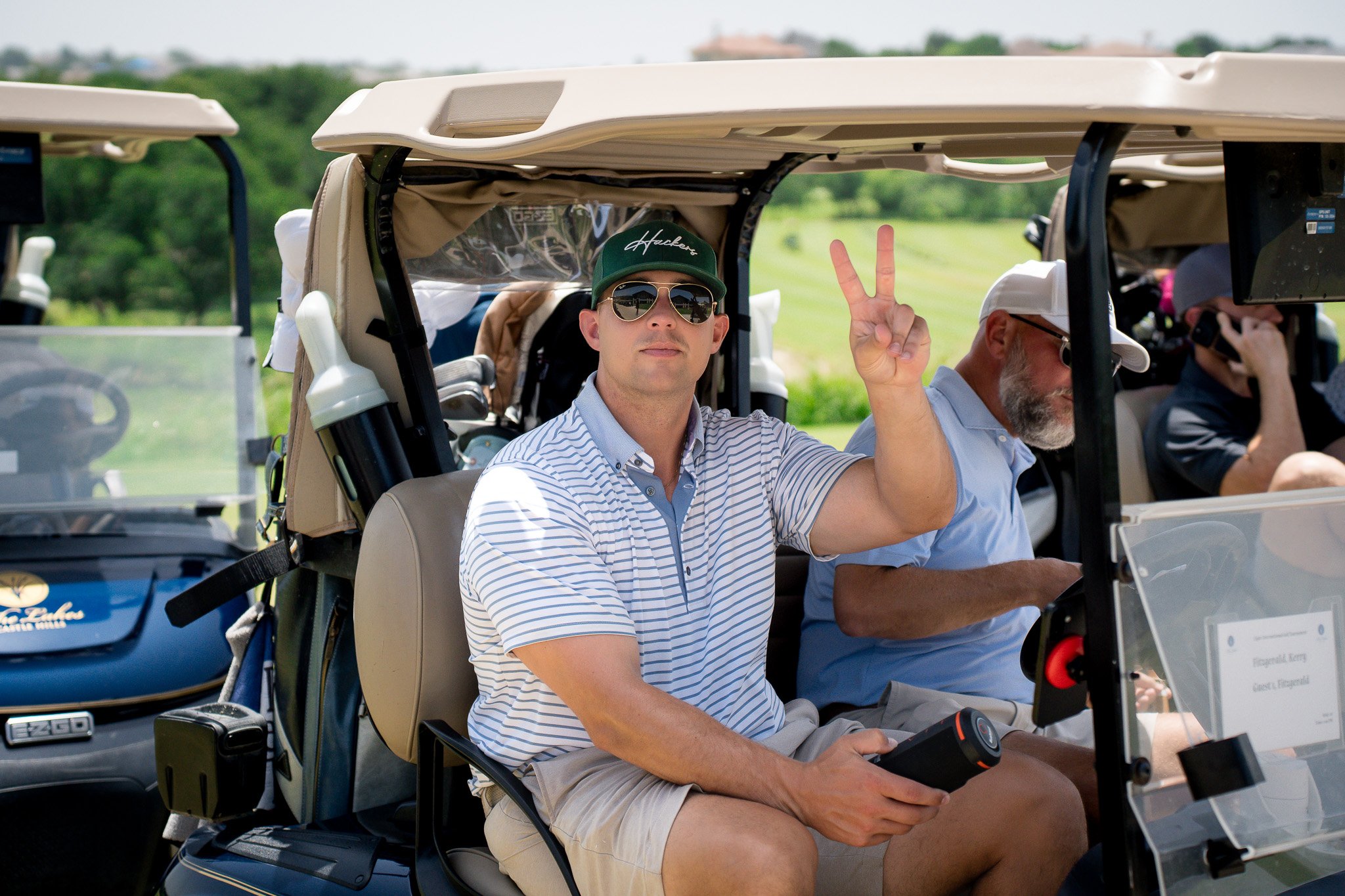 A man wearing sunglasses, a green cap, and a striped polo shirt sitting in a golf cart at a golf course, making a peace sign with his right hand. Other people are also seen in golf carts in the background.
