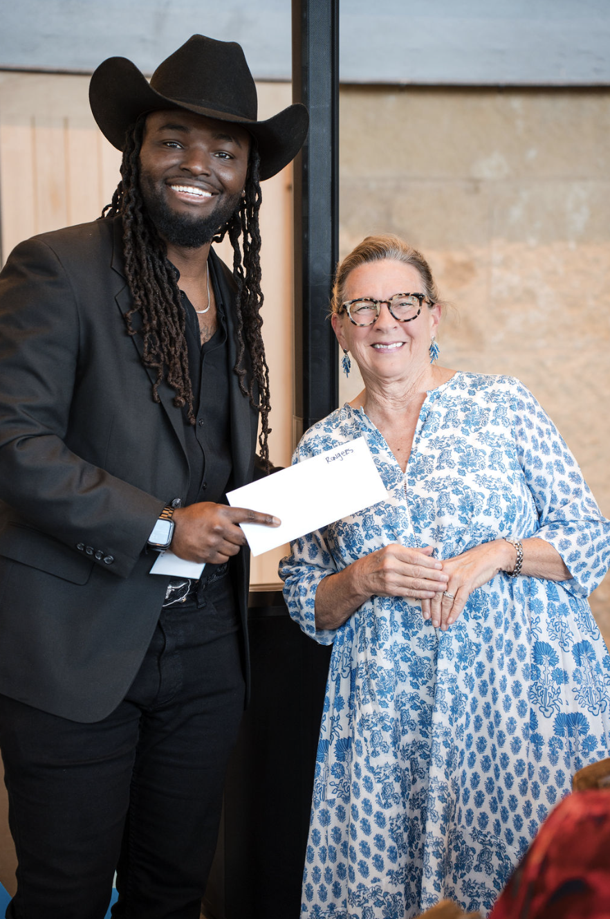 A man with long dreadlocks, wearing a cowboy hat, black suit, and black shirt, is smiling and holding an envelope. Next to him, a woman with short hair, glasses, and blue earrings, is smiling and wearing a blue and white floral dress.