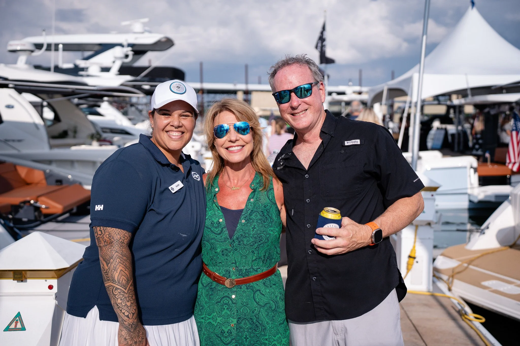 Three smiling people standing together at a marina with boats and yachts in the background. The woman on the left has tattoos on her arm and is wearing a dark polo shirt and a white hat. The woman in the middle has blonde hair, blue sunglasses, and i