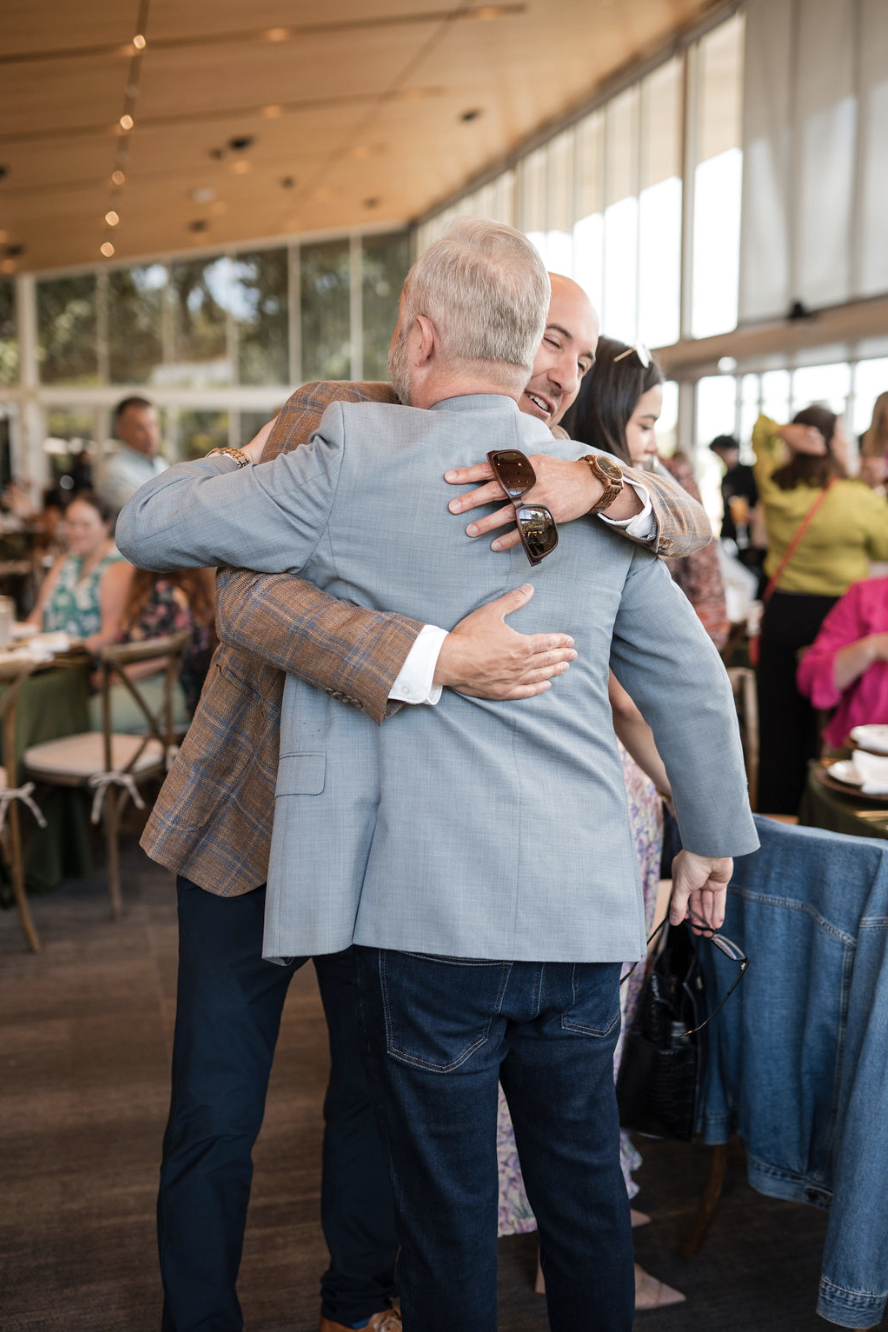 Two men hugging at a gathering in a bright, modern room with large windows, with other people seated and standing in the background.
