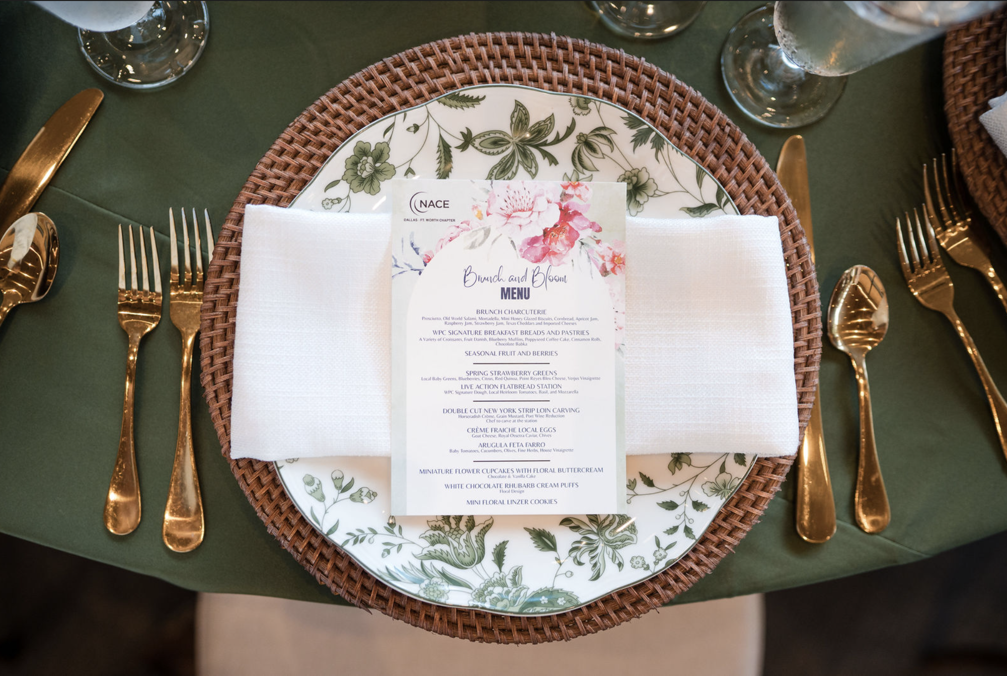 Elegant place setting including a floral patterned plate, gold cutlery, a white cloth napkin, and a floral menu card on a green tablecloth.