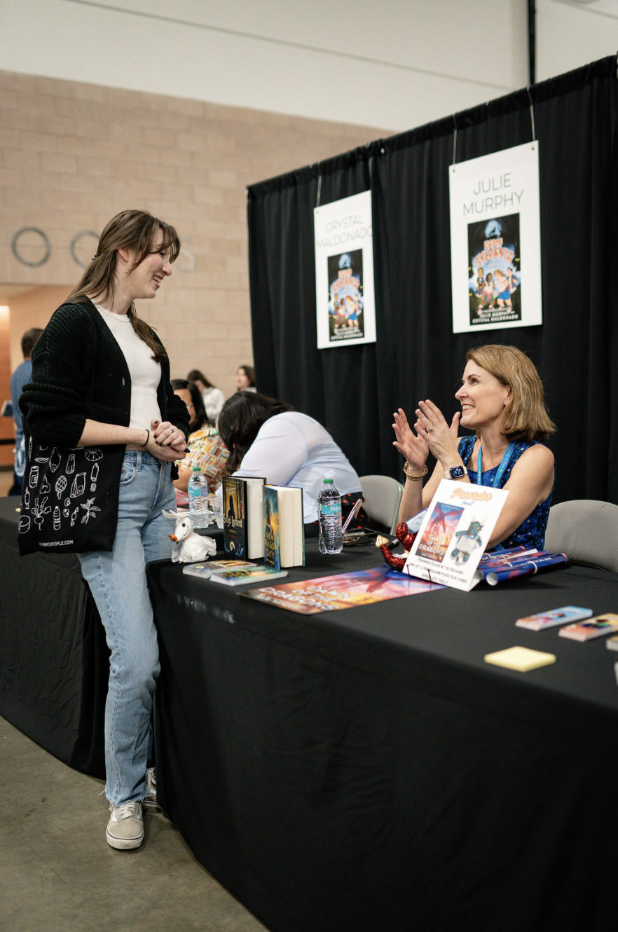 A woman with blonde hair wearing a blue patterned top sitting at a table, smiling and clapping, talking to a young woman with long brown hair wearing a black cardigan, white shirt, and jeans, at a book signing event. The table has books, water bottle