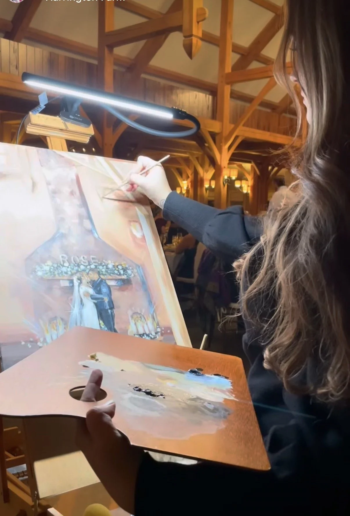 A woman creating a wedding portrait of a bride and groom with the word 'ROSE' during a painting class in a rustic wooden hall.