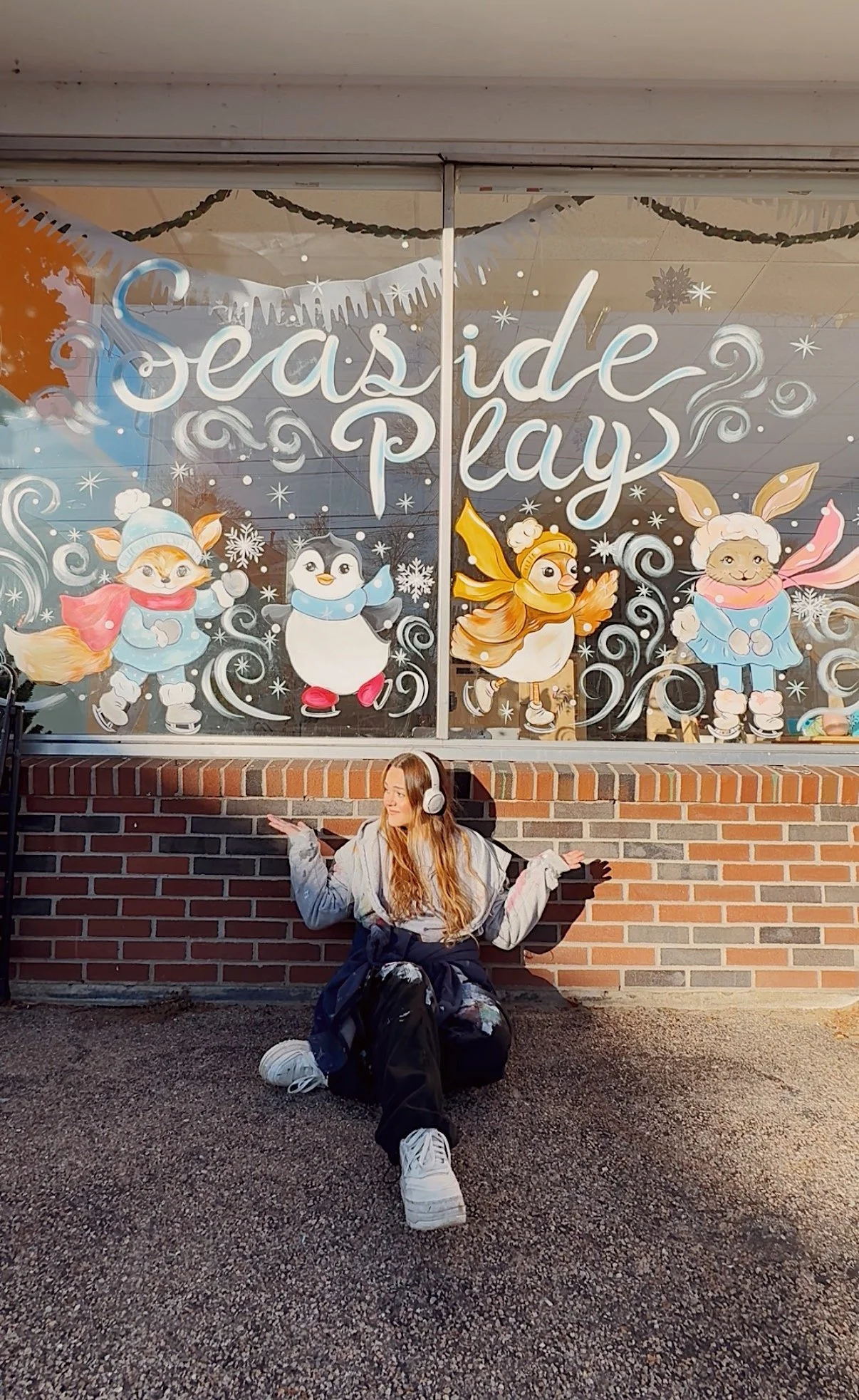A girl sitting on the ground wearing headphones, with a decorative window in the background featuring festive characters and text that reads 'Seaside Play'.