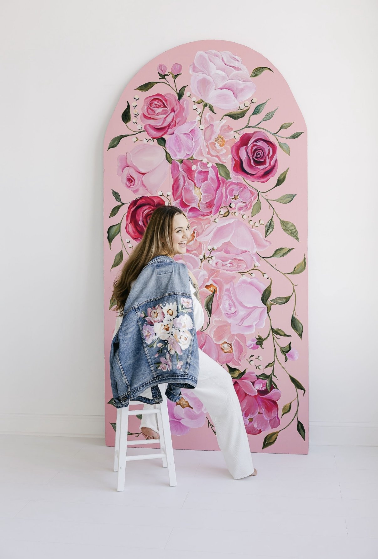 Young woman sitting on a white stool in front of a large pink floral wall mural, smiling and looking over her shoulder.