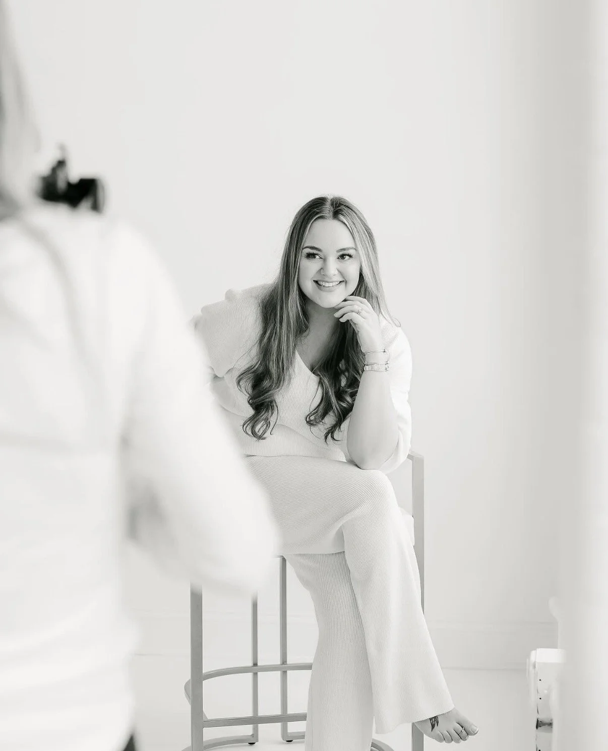 A woman sitting on a stool, smiling and posing for a photo in a minimalistic photo studio.