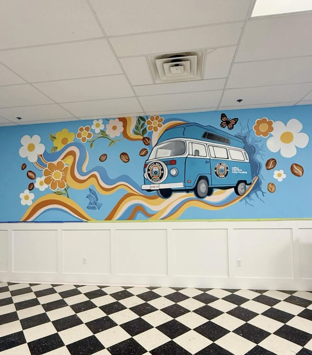 Colorful mural of a blue camper van surrounded by flowers, coffee beans, and butterflies painted on an indoor wall with a checkered black and white floor.