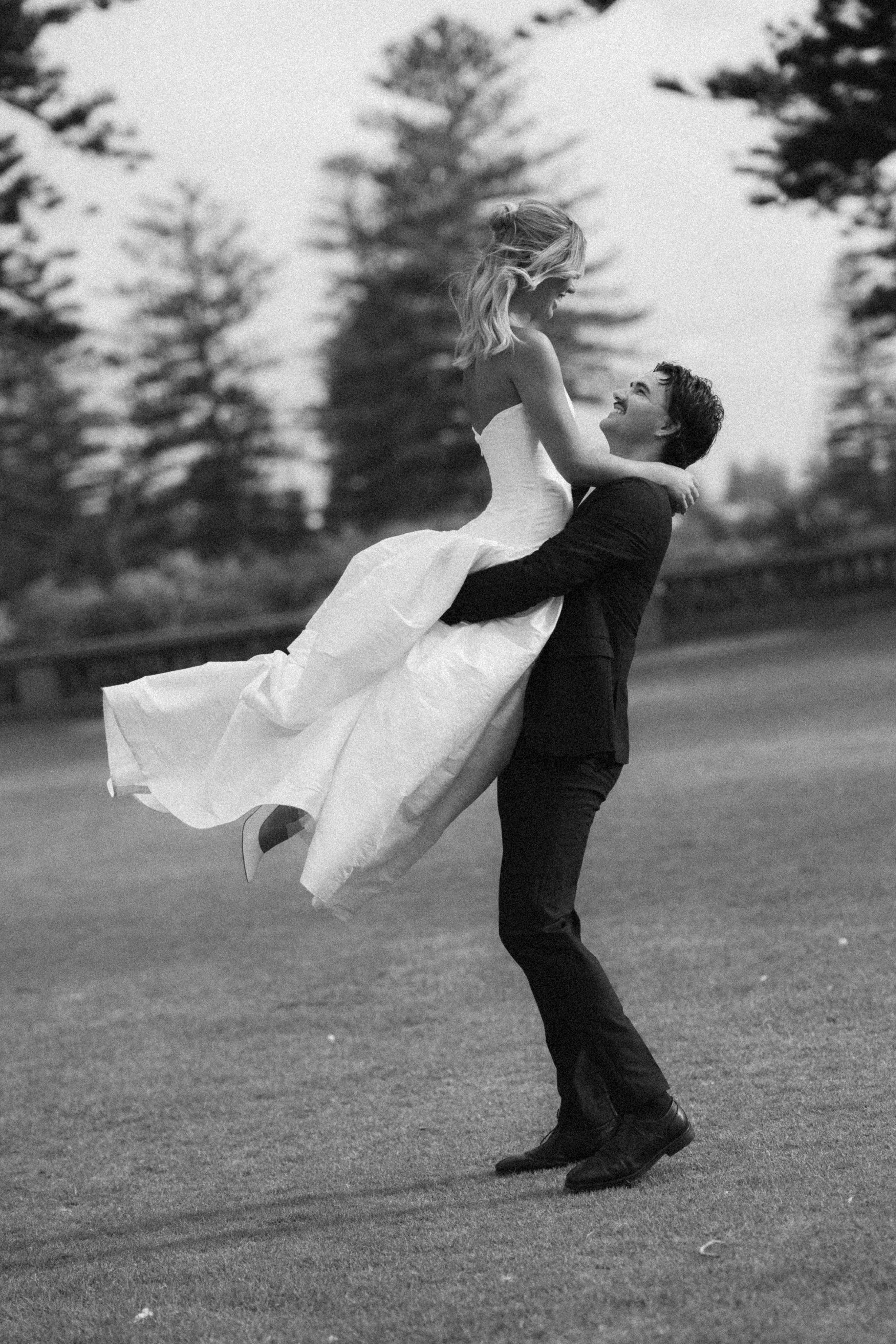 A man in a suit lifting a woman in a wedding dress in an outdoor setting with trees in the background, black and white photograph.