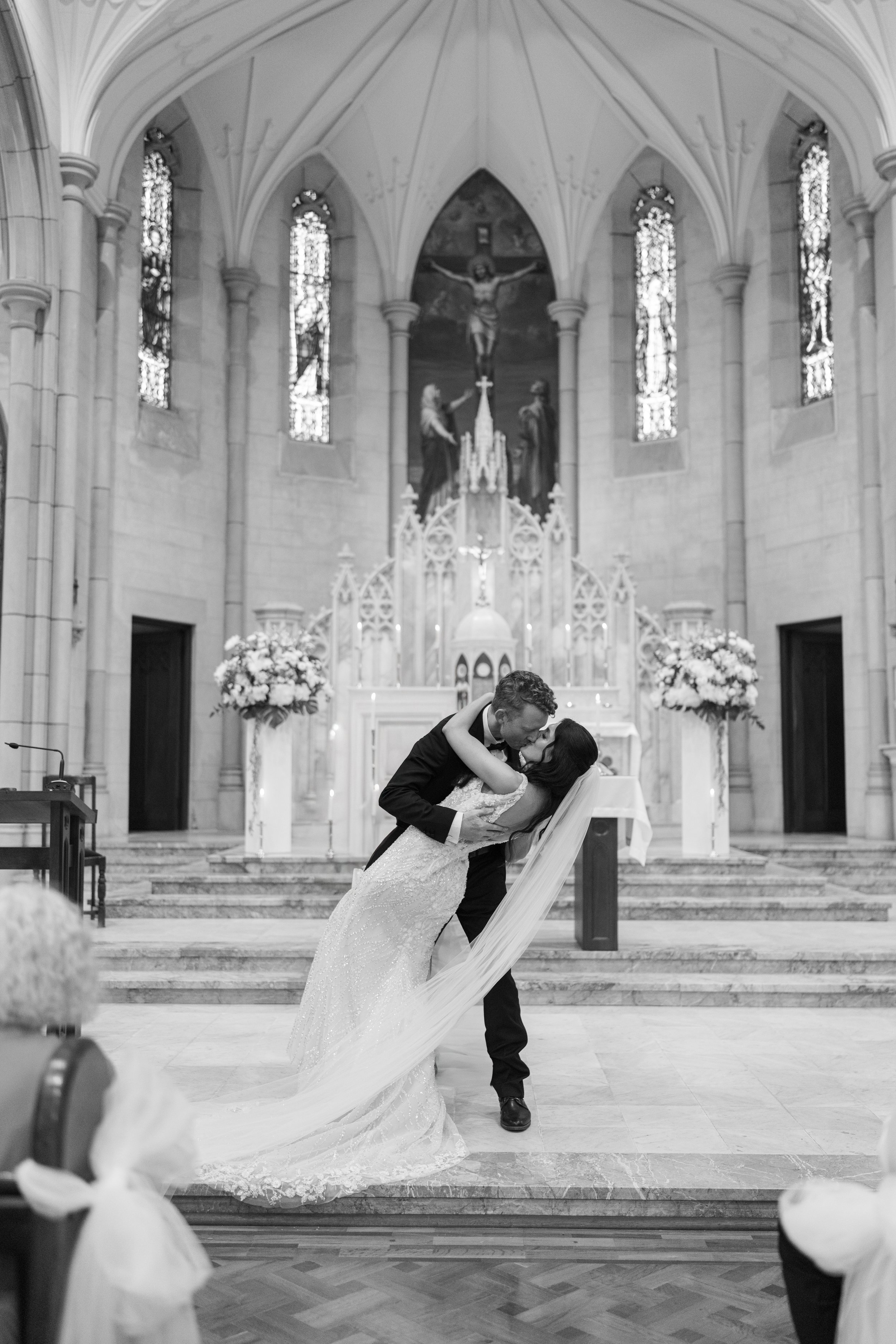 A newlywed couple sharing a kiss inside a church altar with gothic architecture, stained glass windows, and floral arrangements.