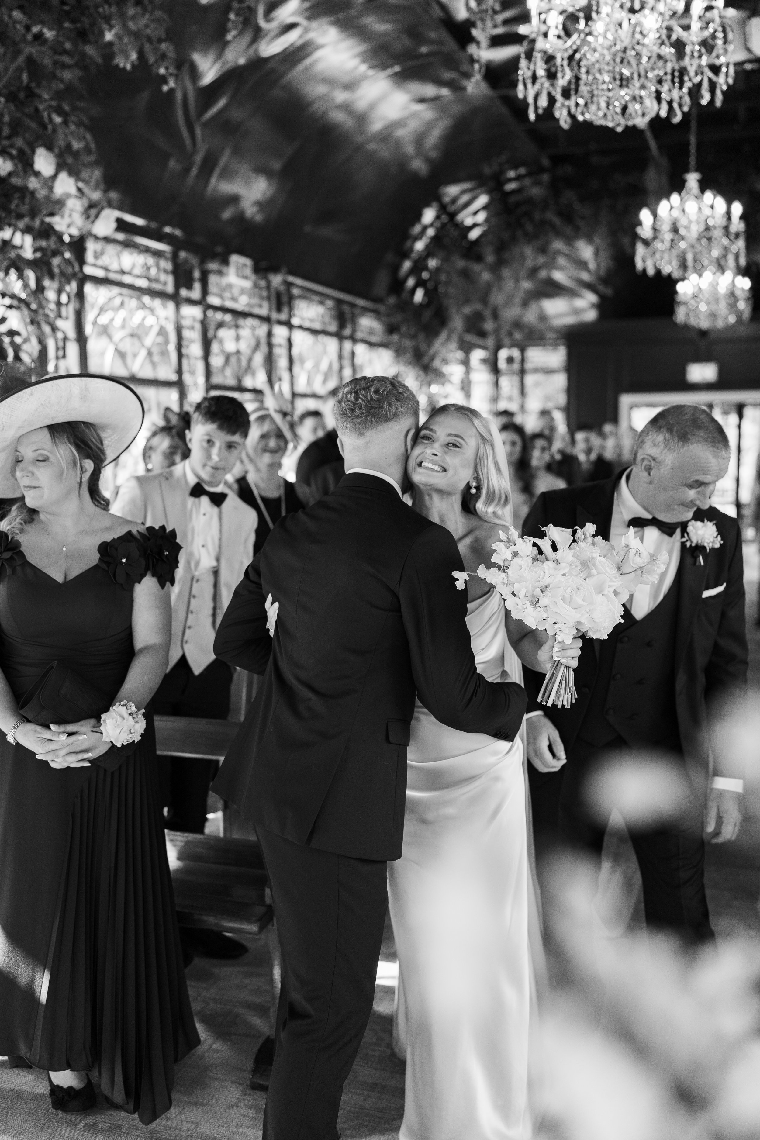 Black and white photograph of a wedding ceremony with a bride in a satin gown holding a bouquet, hugging a groom in a tuxedo, surrounded by family and guests.