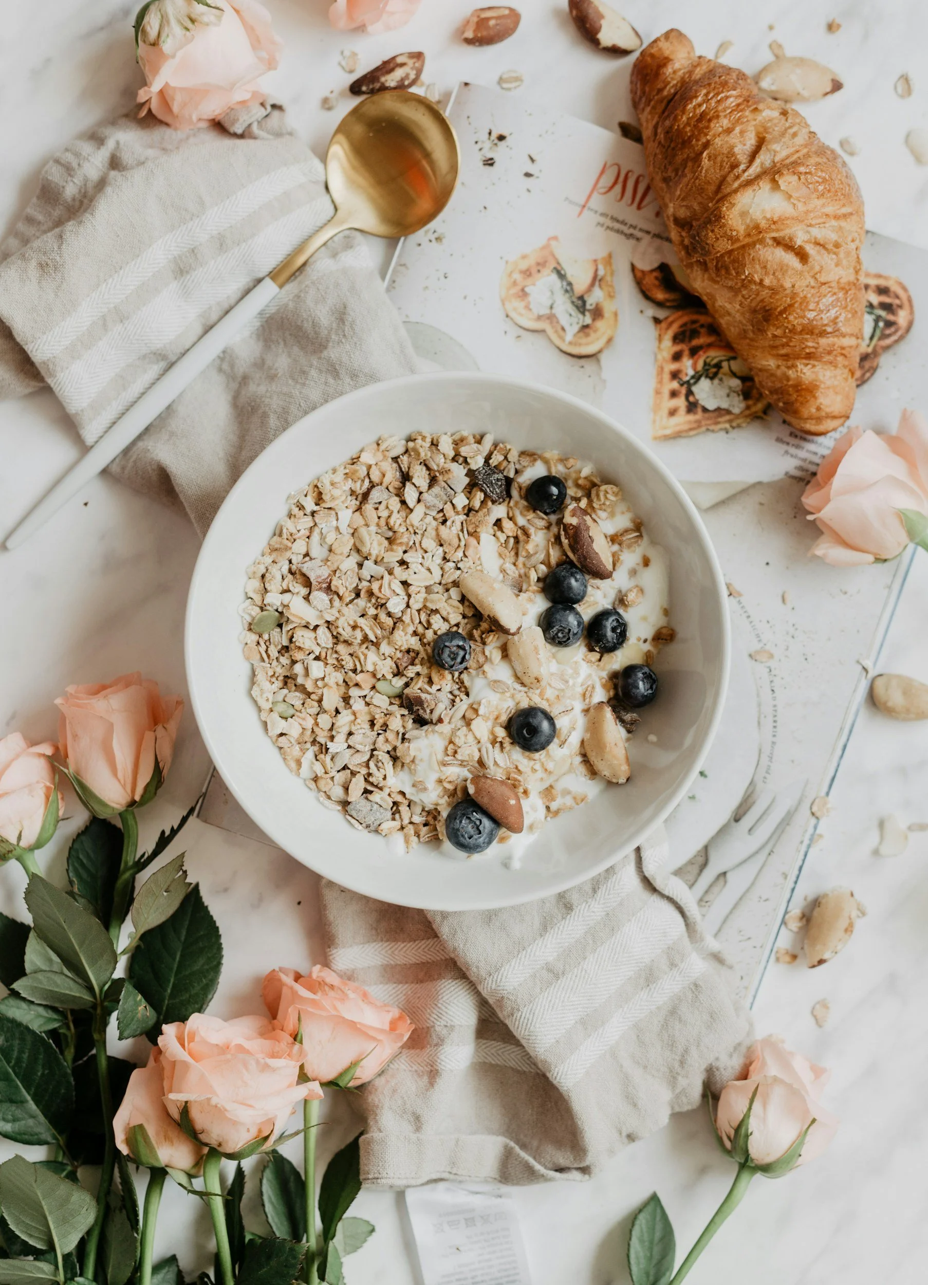 A breakfast scene with a bowl of yogurt topped with granola, blueberries, and nuts, placed on a cloth napkin. Surrounding are pink roses, a croissant, a gold spoon, and scattered almonds on a white surface.