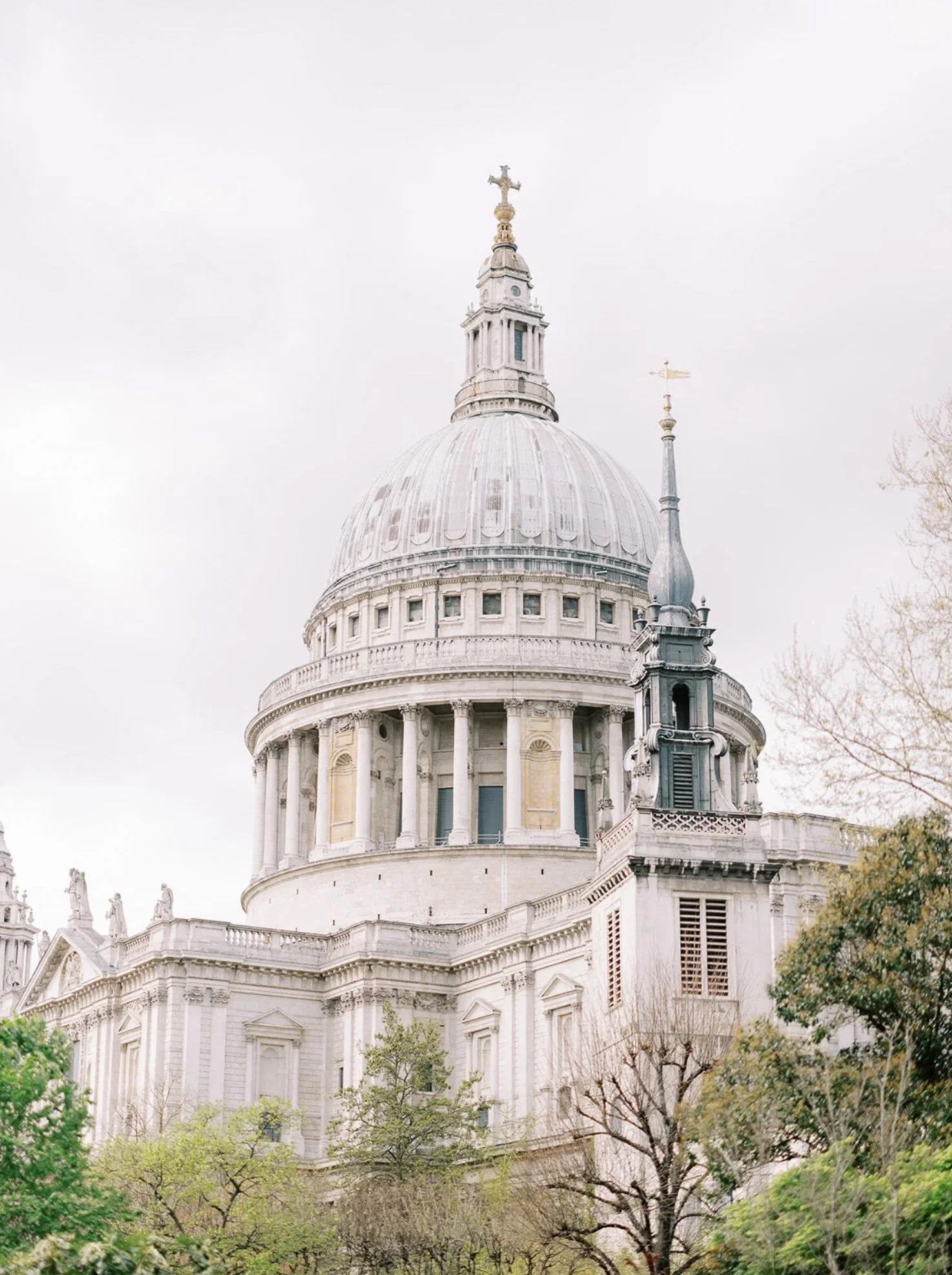 The image shows the dome of St. Paul's Cathedral in London, UK, with a cross on top and surrounding architectural details, partially obscured by trees.