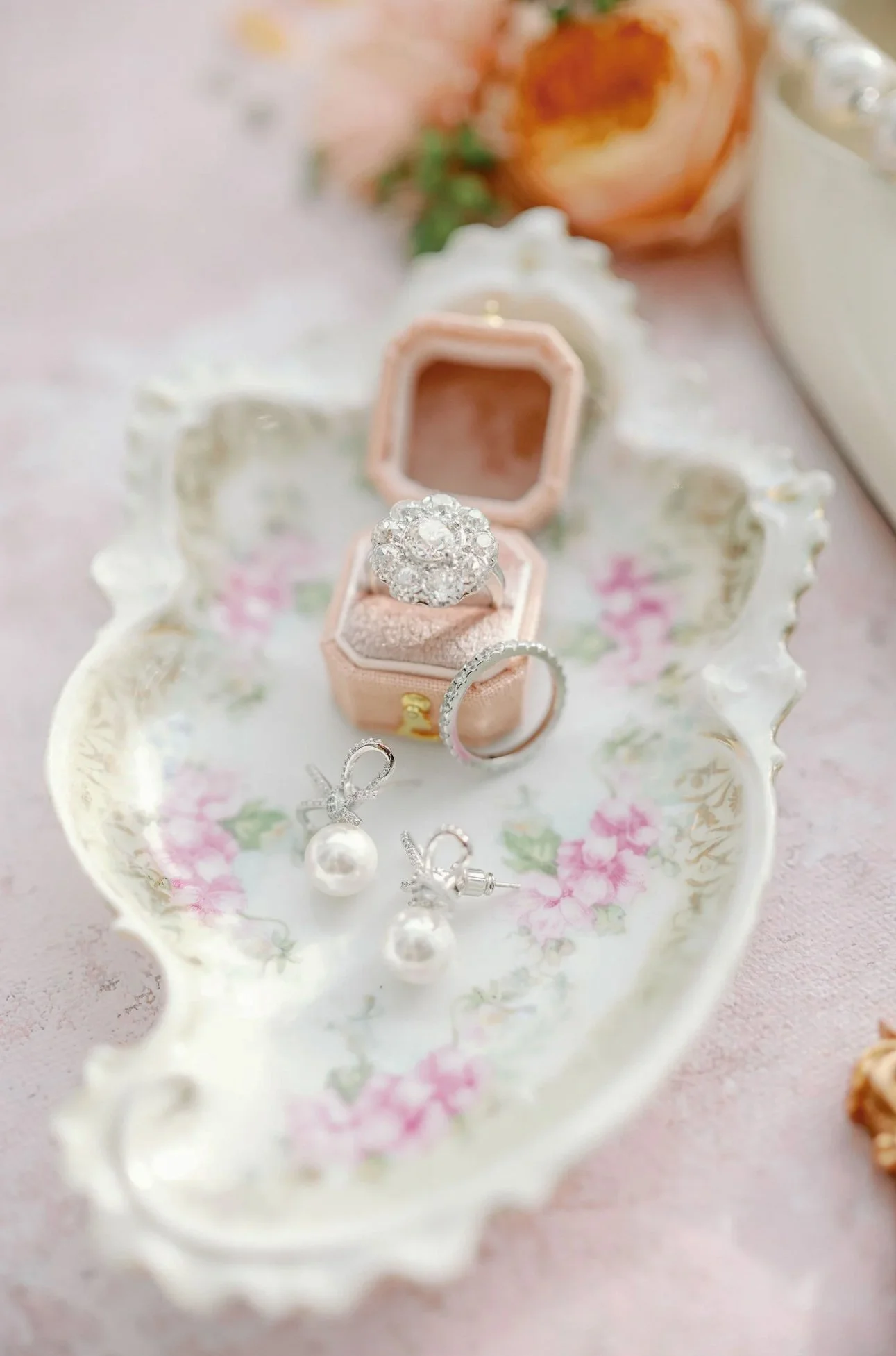 A jewelry display with sparkling diamond rings in a pink velvet ring box and pearl earrings on a floral porcelain tray, with a blurred background.