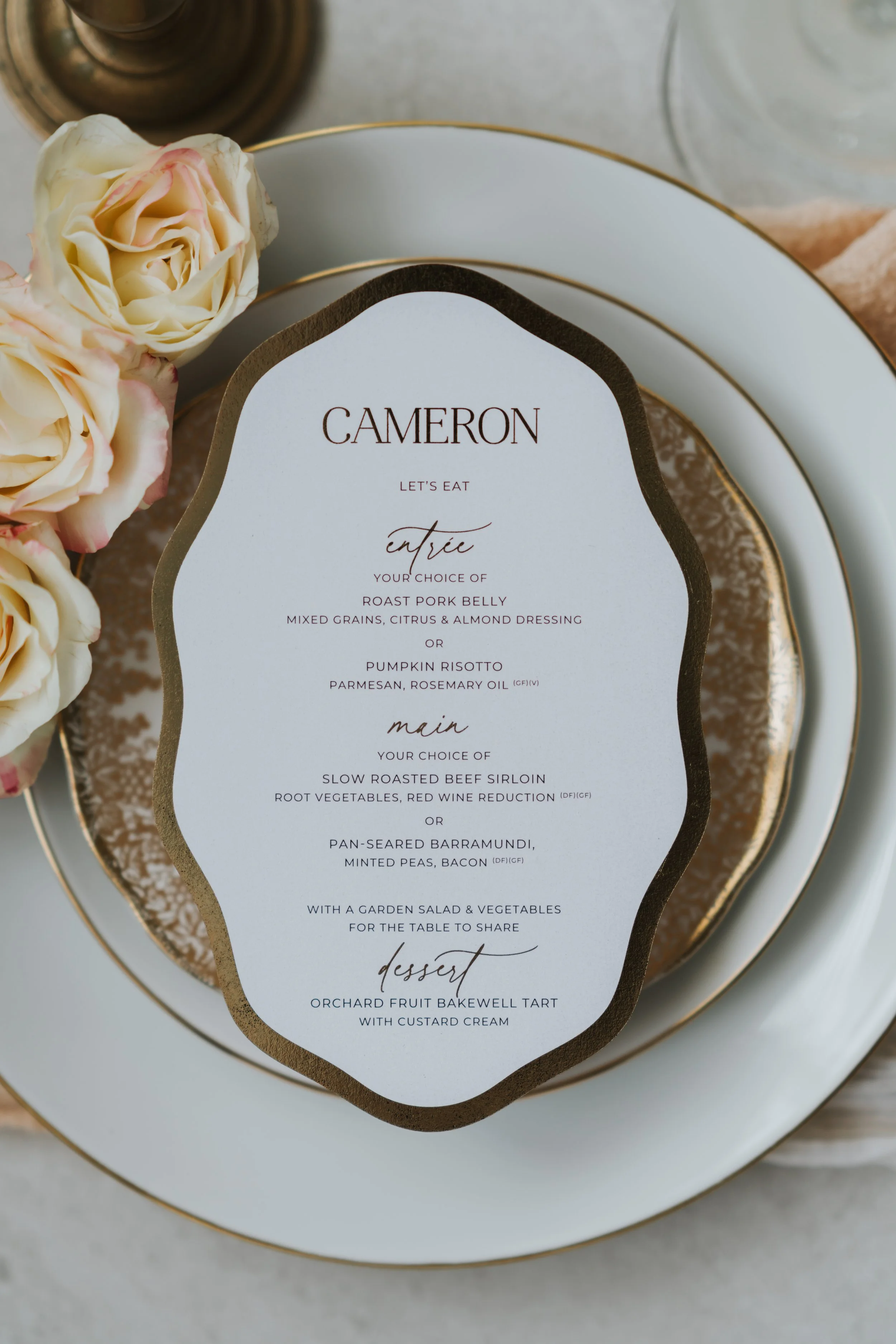 Elegant printed menu placed on a plate with pink roses nearby, featuring a wedding or event meal with choices for starters, mains, and dessert, including details about ingredients and accommodations for dietary preferences.