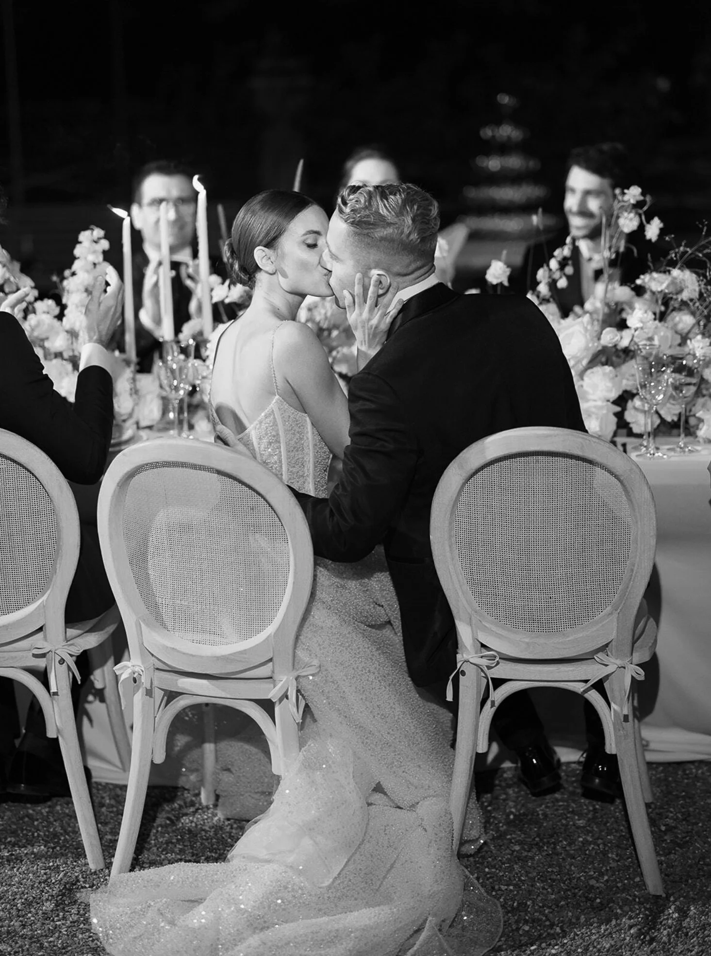 A black and white photograph of a wedding reception, showing a bride and groom kissing at the head table. The bride is wearing a strapless gown, and the groom is in a tuxedo. They are seated on chairs with caned backs, and other guests are visible in the background, along with floral arrangements and lit candles on the table.