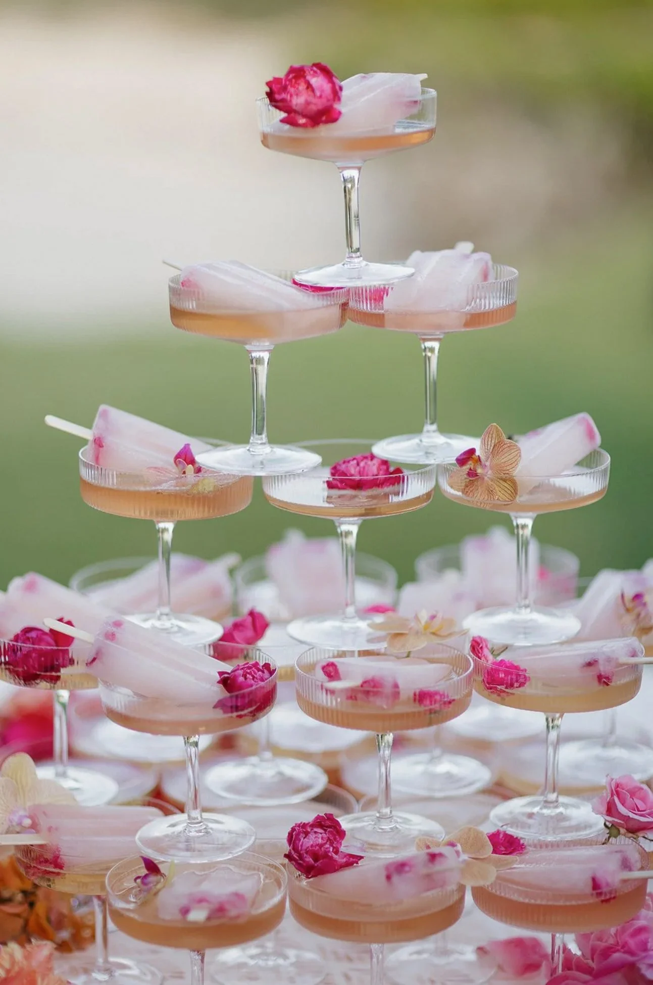 A pyramid of champagne glasses filled with pink cocktails, garnished with edible flowers and flower petals.